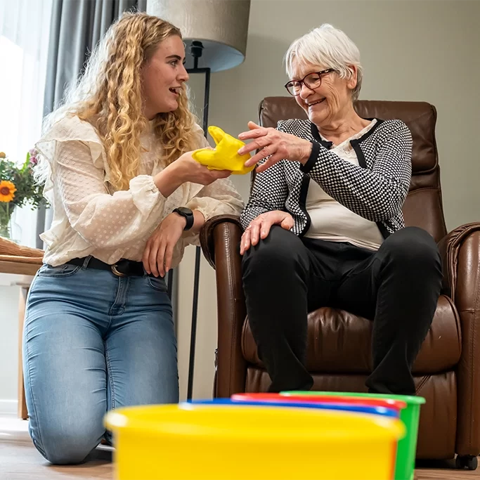 Jonge vrouw begeleidt oudere vrouw met een geel oefenobject in de woonkamer.