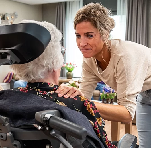 Verzorgende legt geruststellende hand op schouder van oudere in rolstoel in huiskamer.