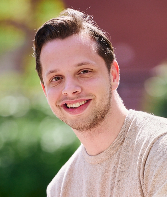 Lachende jongeman met kort haar in beige trui buiten, met groene, zonnige achtergrond.