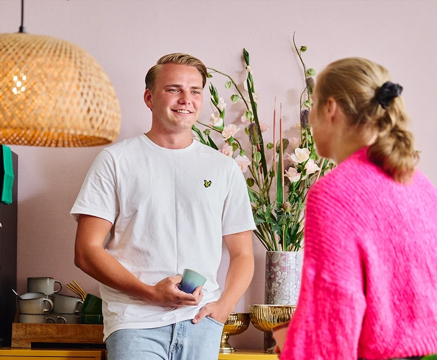 Twee mensen praten ontspannen in een licht interieur; man met mok in wit T-shirt, vrouw in roze trui.