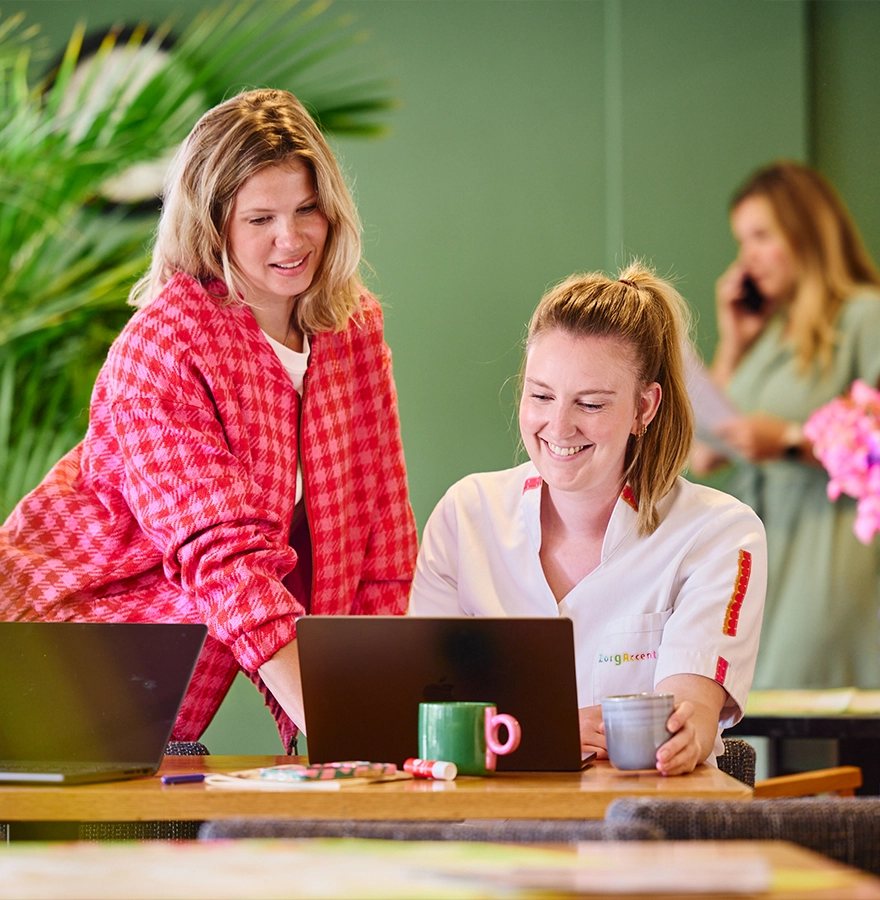 Twee collega’s werken lachend aan laptops in een kleurrijke kantoorruimte met planten; op de achtergrond belt iemand.