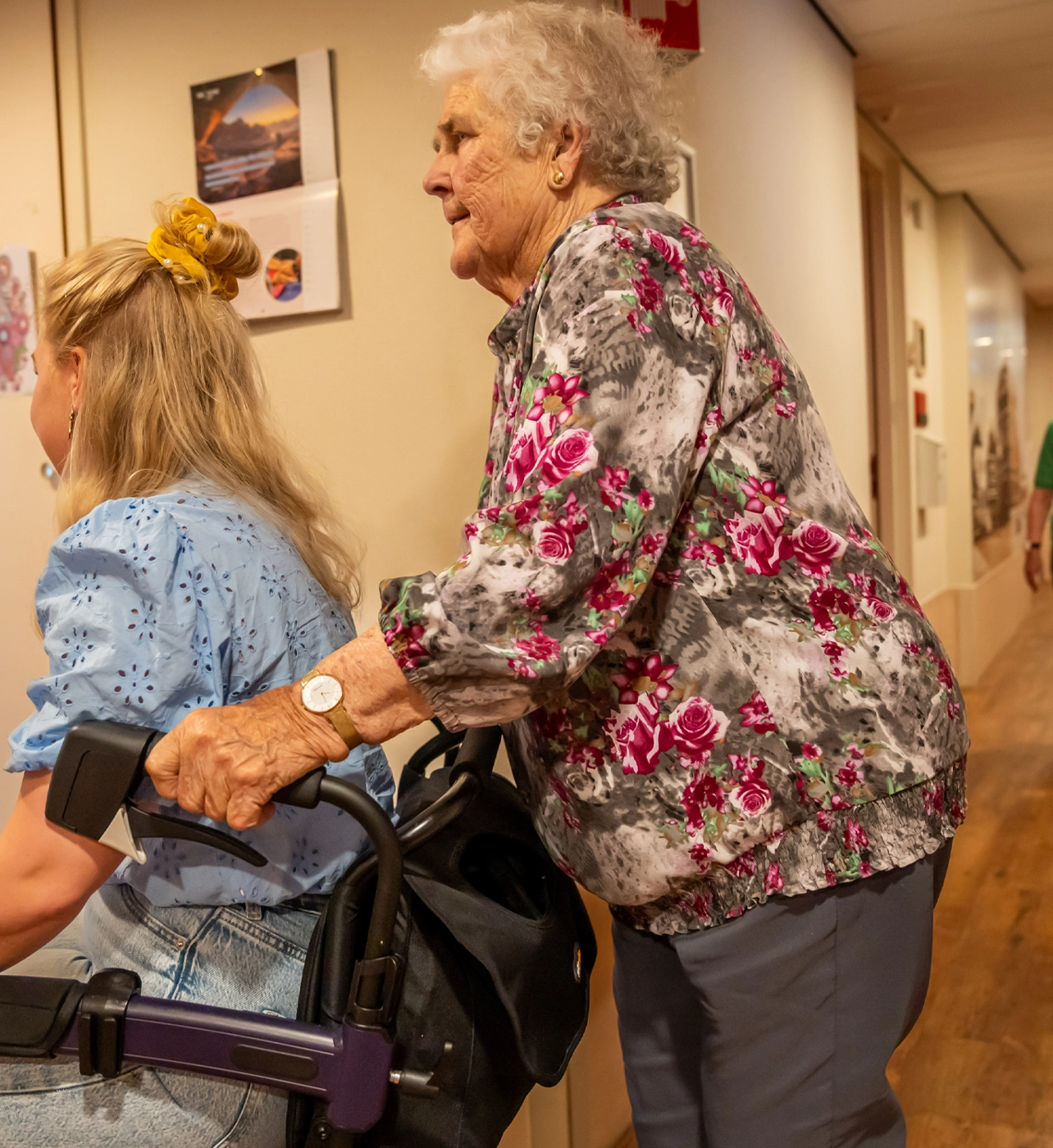 Oudere vrouw met bloemetjesblouse duwt rollator in een gang, naast jongere vrouw.