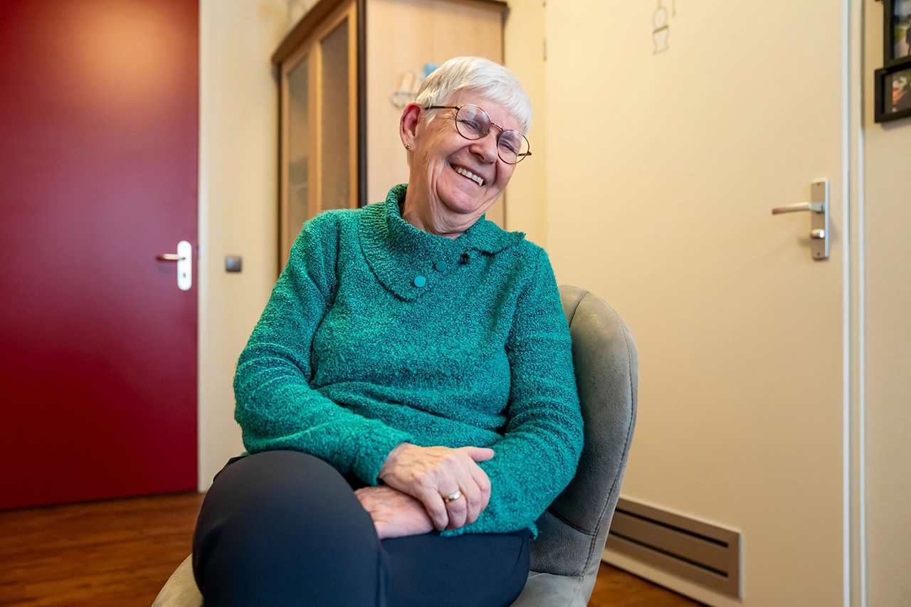 Lachende oudere vrouw met bril in groene trui, zittend in een stoel in huiselijke kamer.