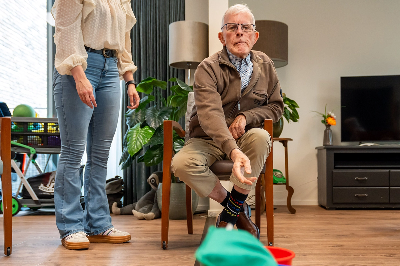 Oudere man gooit een bal tijdens een oefenspel in de huiskamer, begeleid door een vrouw.
