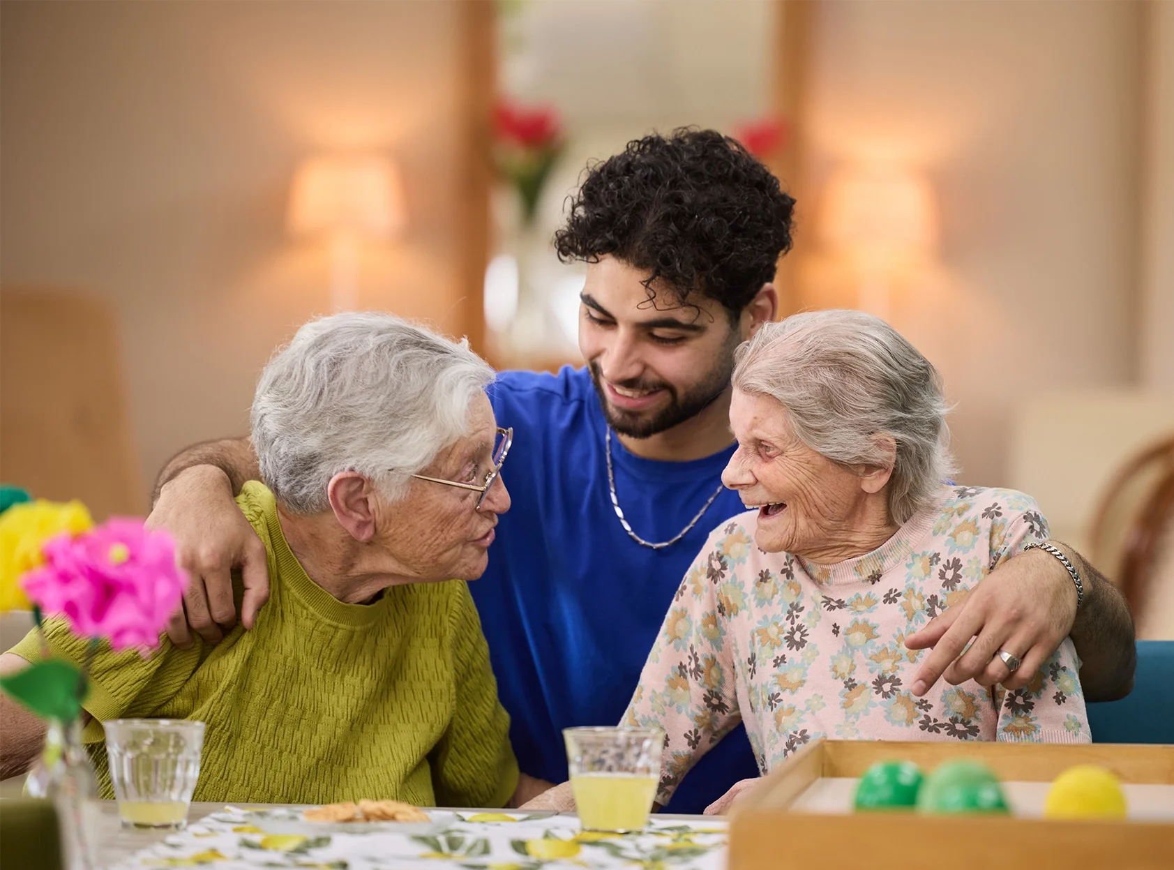Jonge zorgmedewerker omarmt twee lachende ouderen aan tafel in een warme zorginstelling.