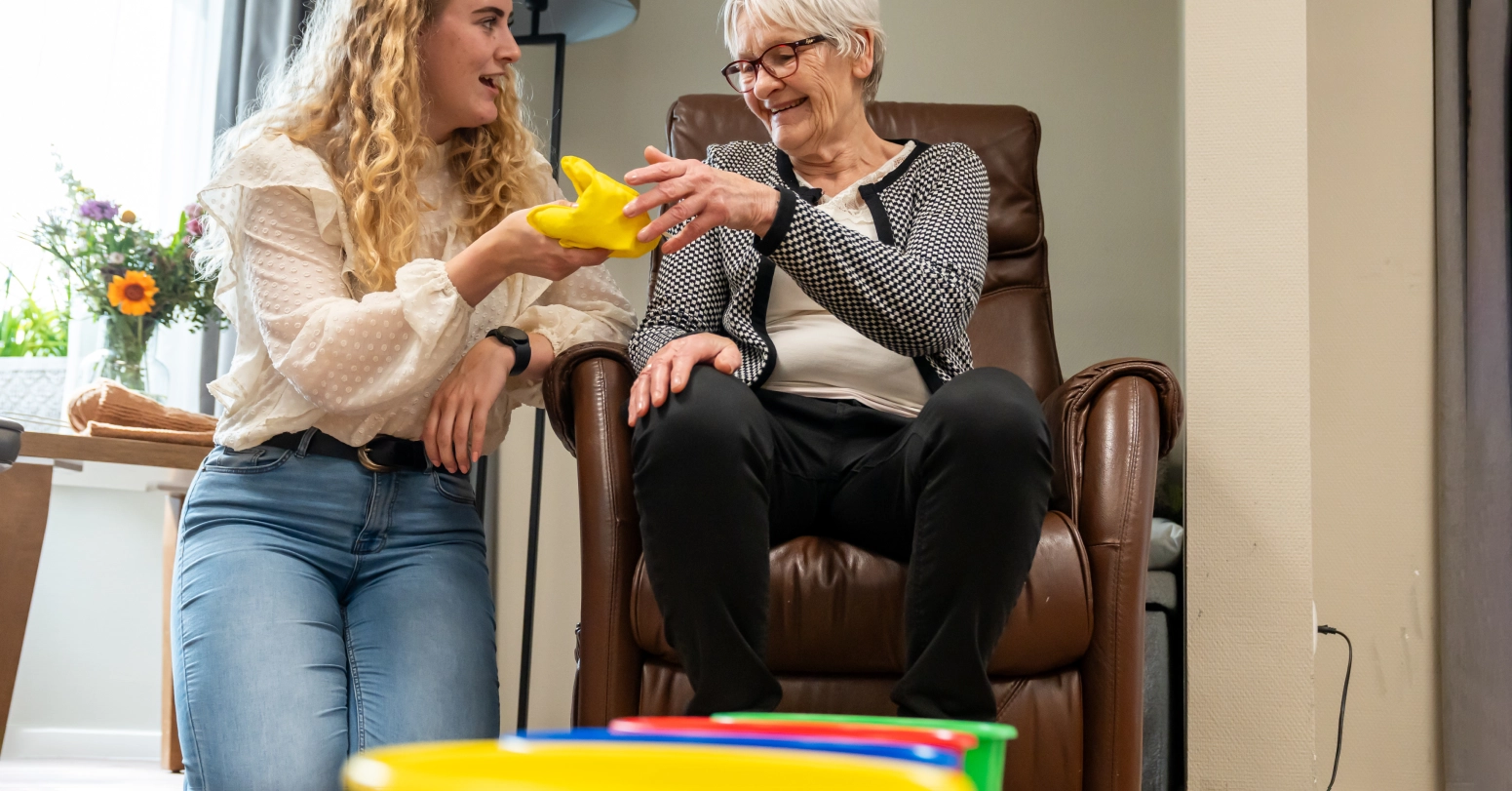 Jonge vrouw naast oudere vrouw in leunstoel, geeft haar een geel hulpmiddel; beiden glimlachen in de woonkamer