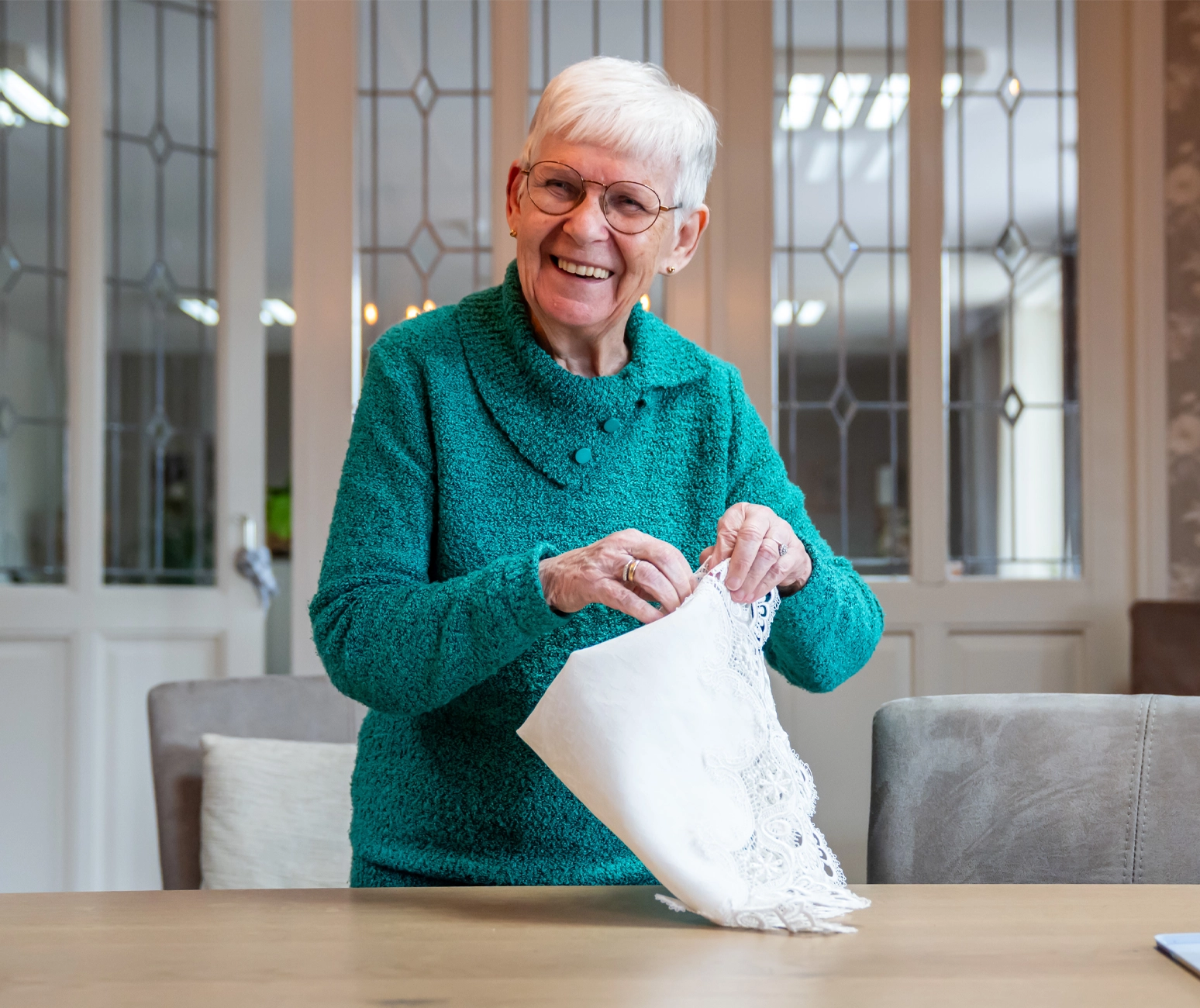 Lachende oudere vrouw vouwt een wit kanten kleed aan een houten tafel thuis.