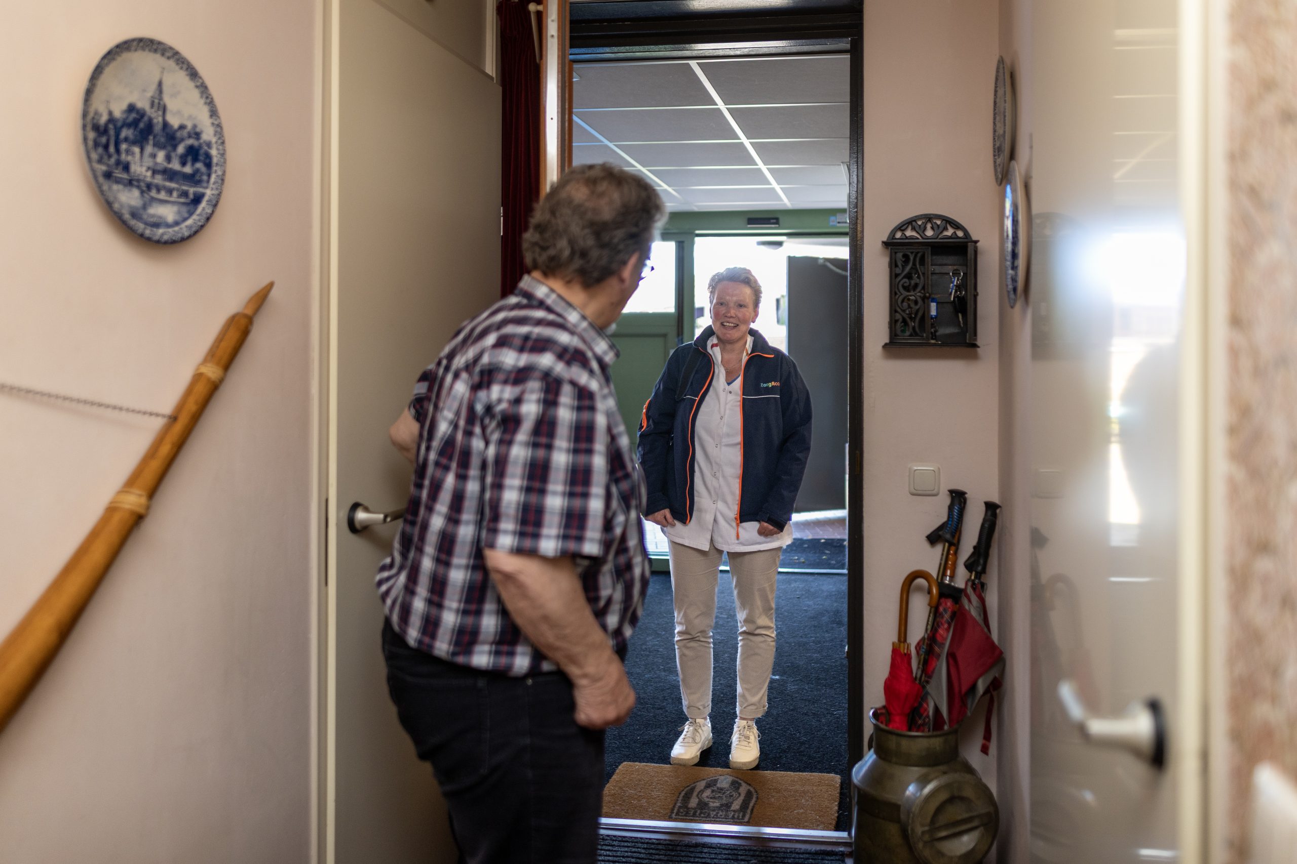 Man opent de voordeur en verwelkomt een lachende vrouw in de hal; paraplubak en deurmat zichtbaar.