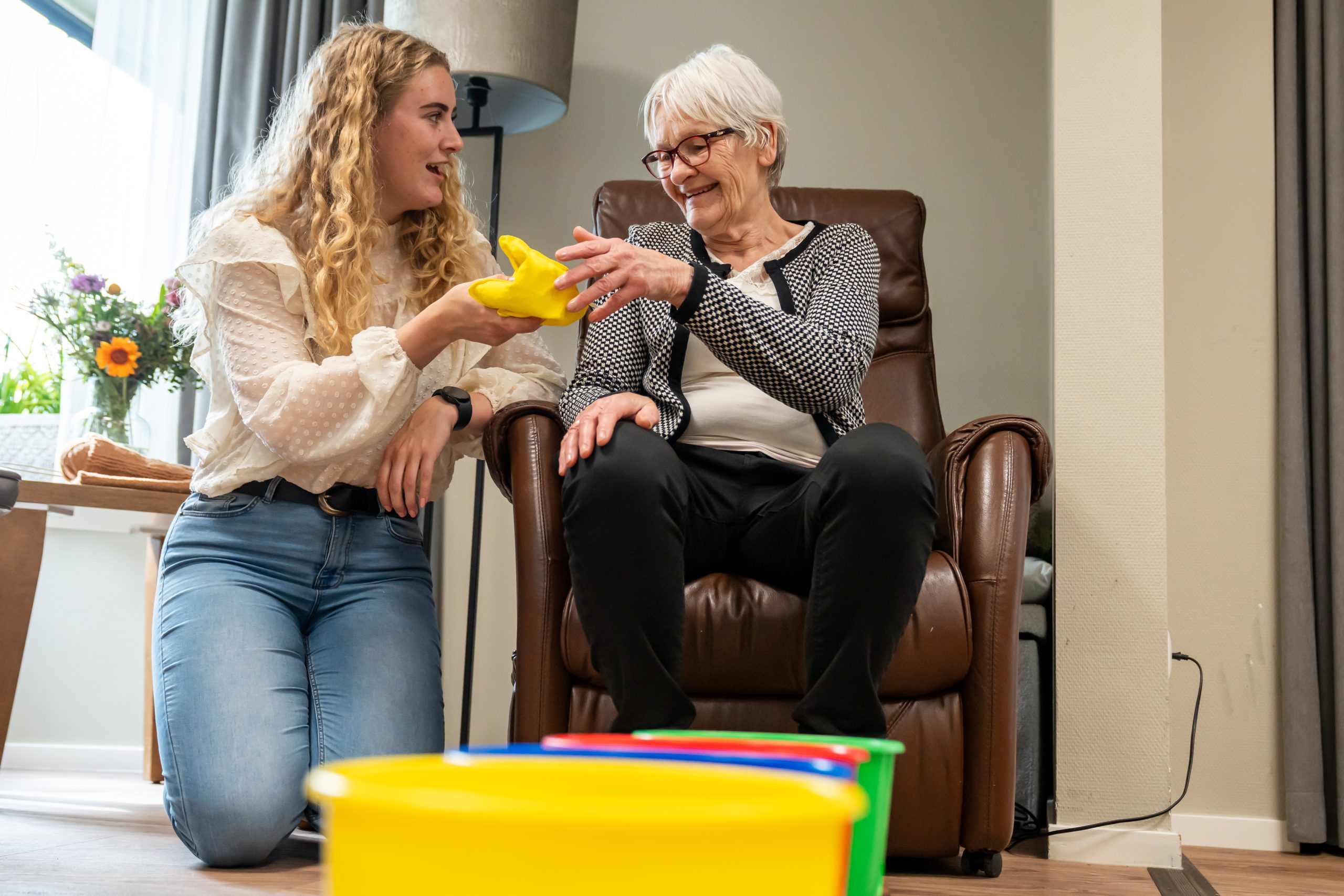 Jonge vrouw helpt oudere vrouw in stoel en geeft haar een geel object in de woonkamer.