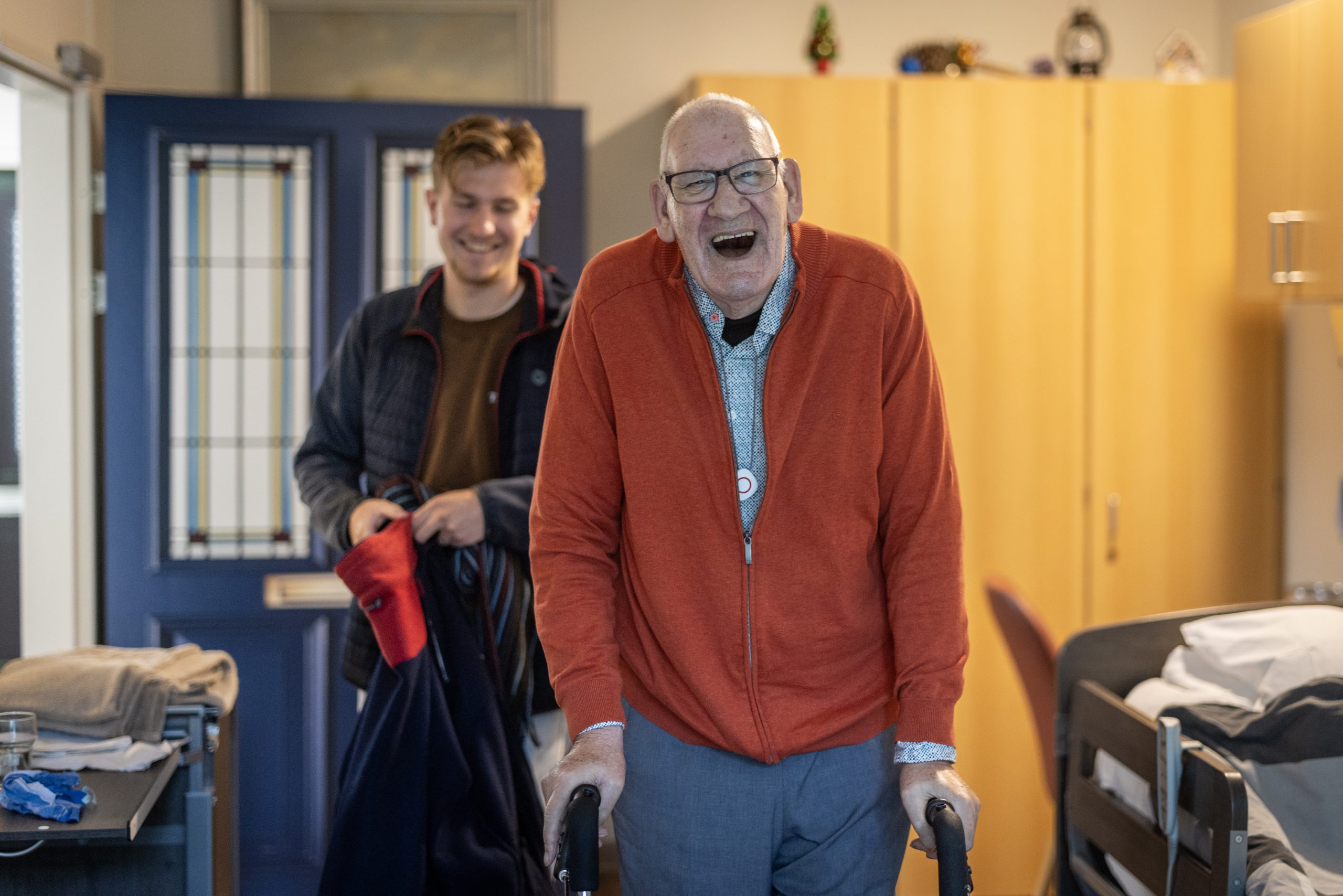 Lachende oudere man met rollator, met jonge begeleider erachter in een zorgkamer.