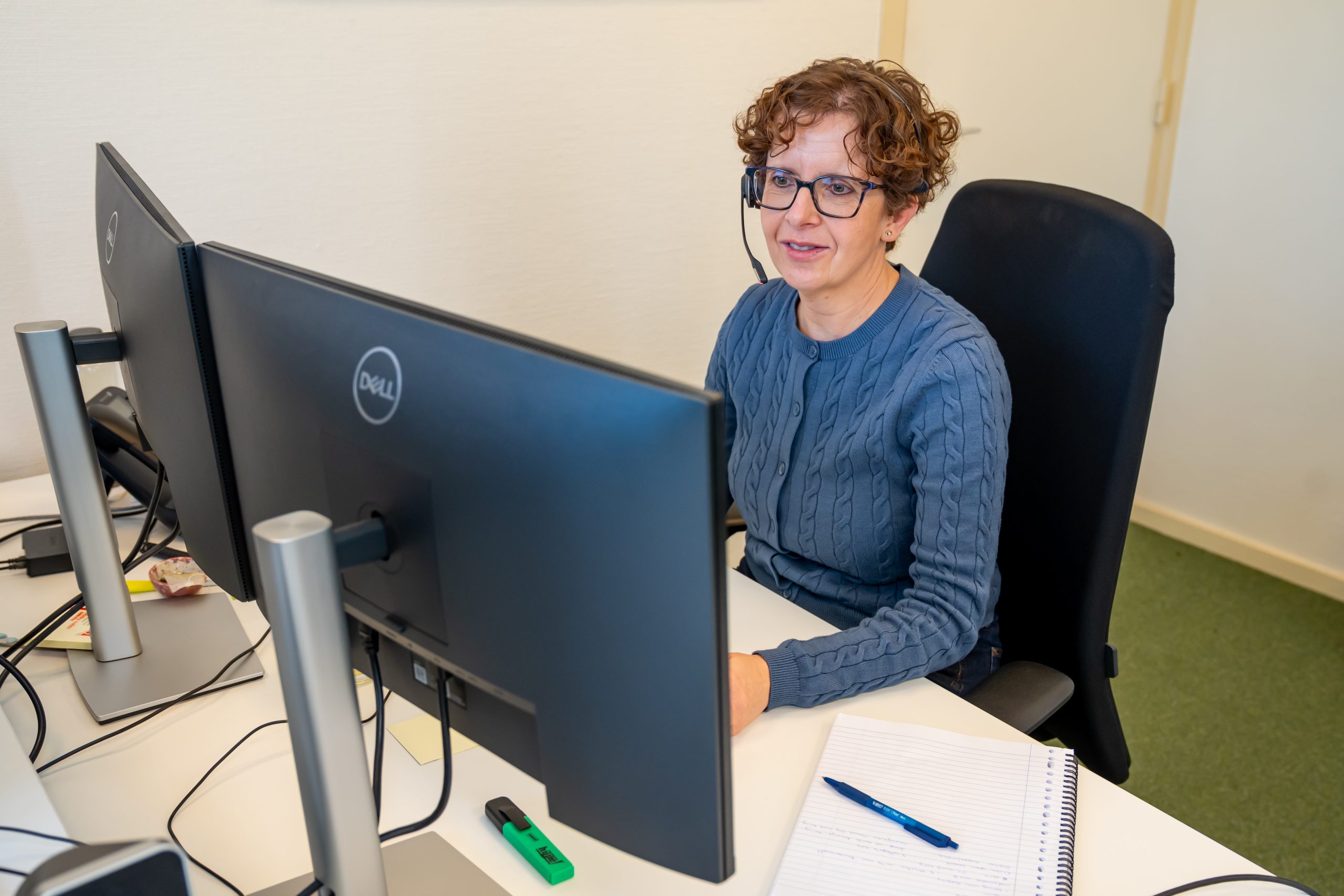 Medewerker met headset werkt achter twee beeldschermen, met notitieboek en pen op een bureau.