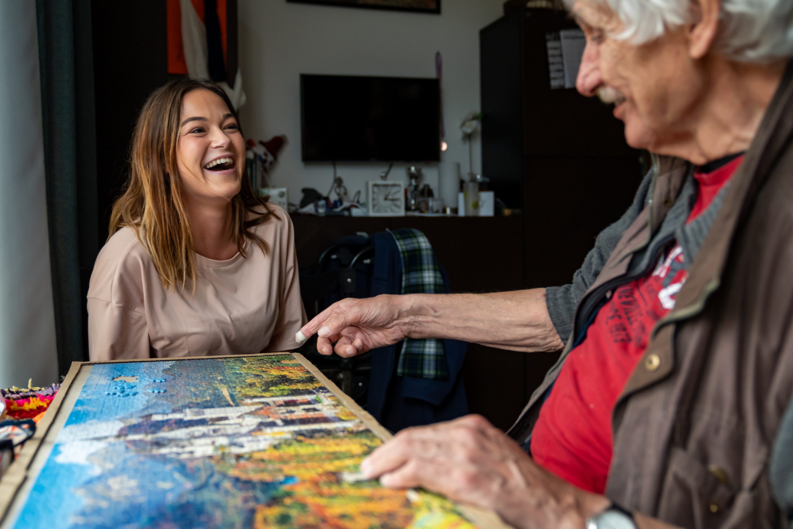 Jonge vrouw lacht terwijl ze met een oudere man samen aan een kleurrijke legpuzzel werkt.