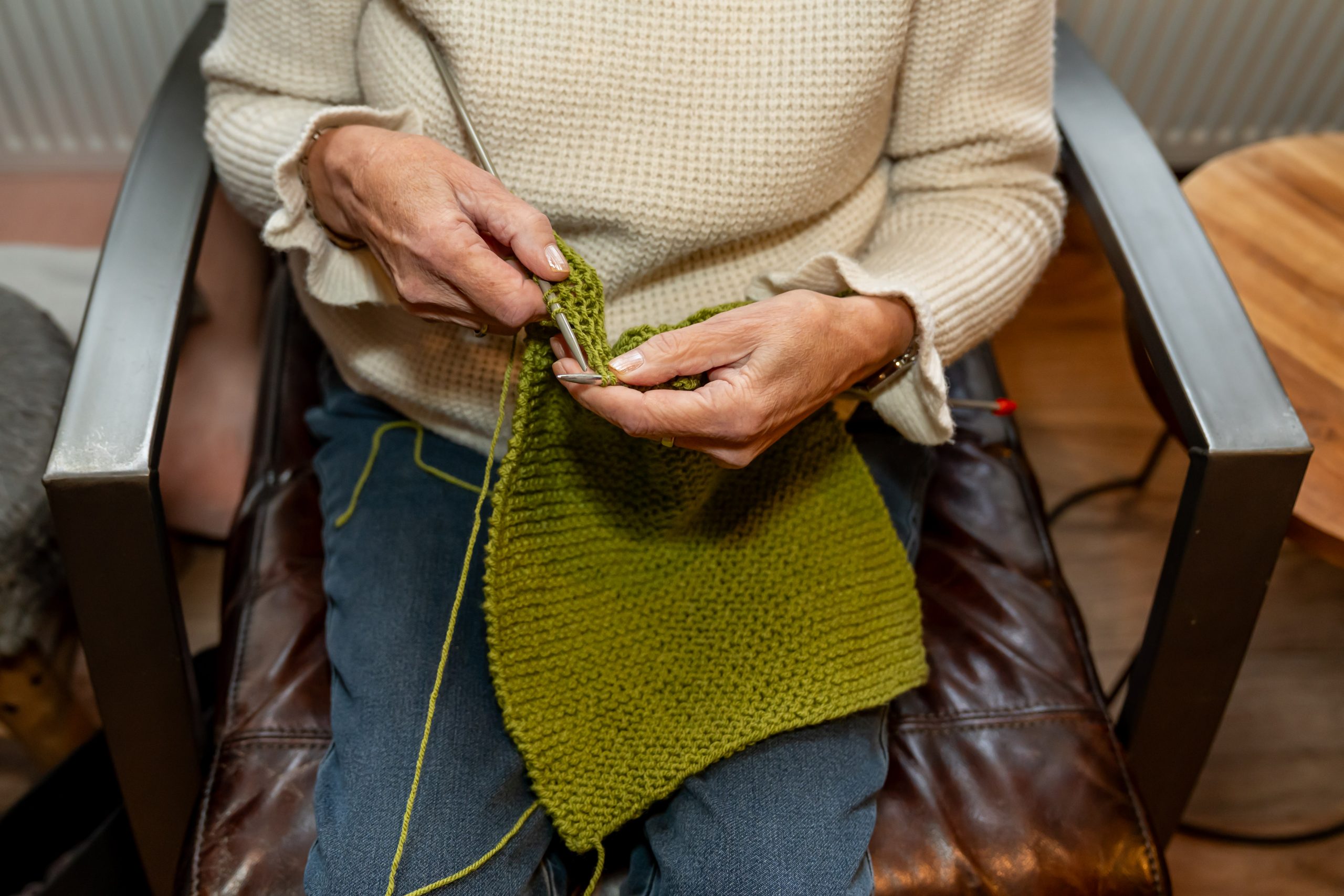 Handen van een persoon die een groene sjaal breit met breinaalden op een stoel.