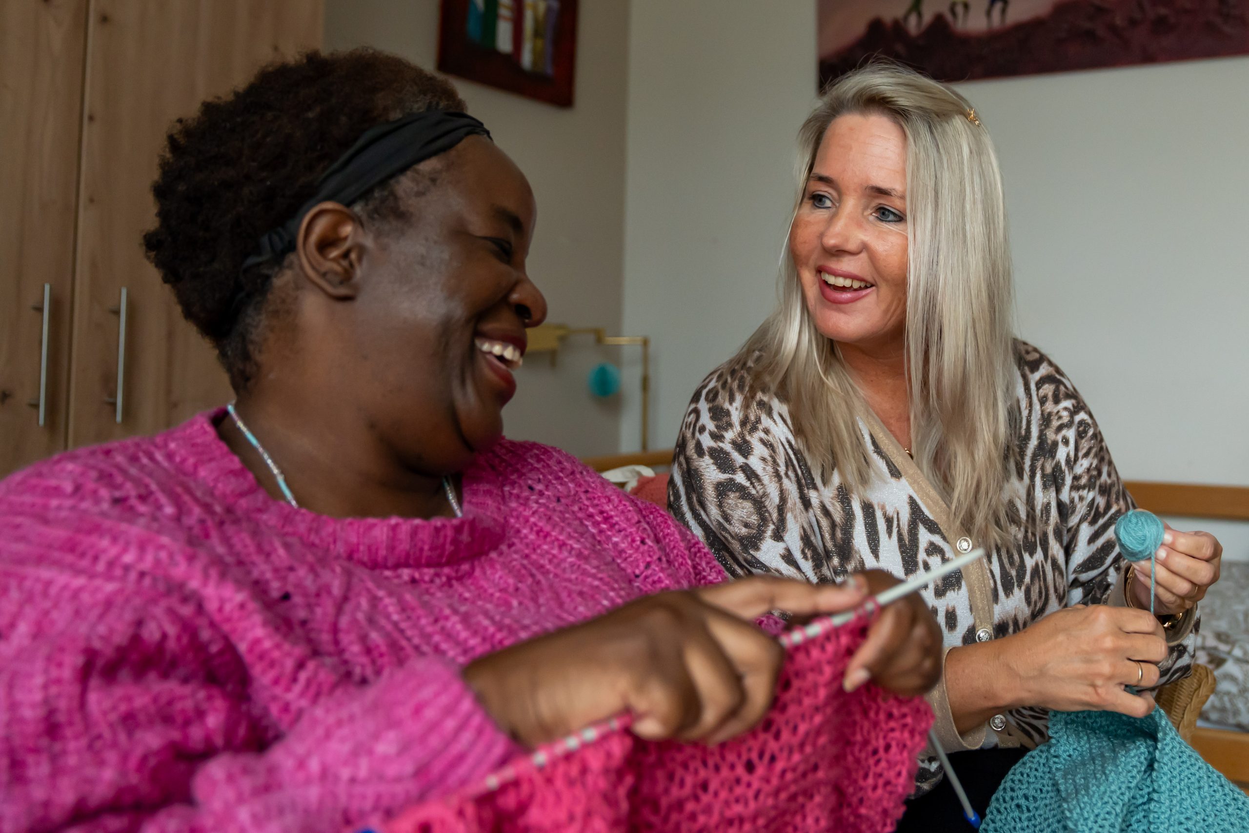 Twee vrouwen lachen terwijl ze samen breien, met roze en turquoise wol op de bank.