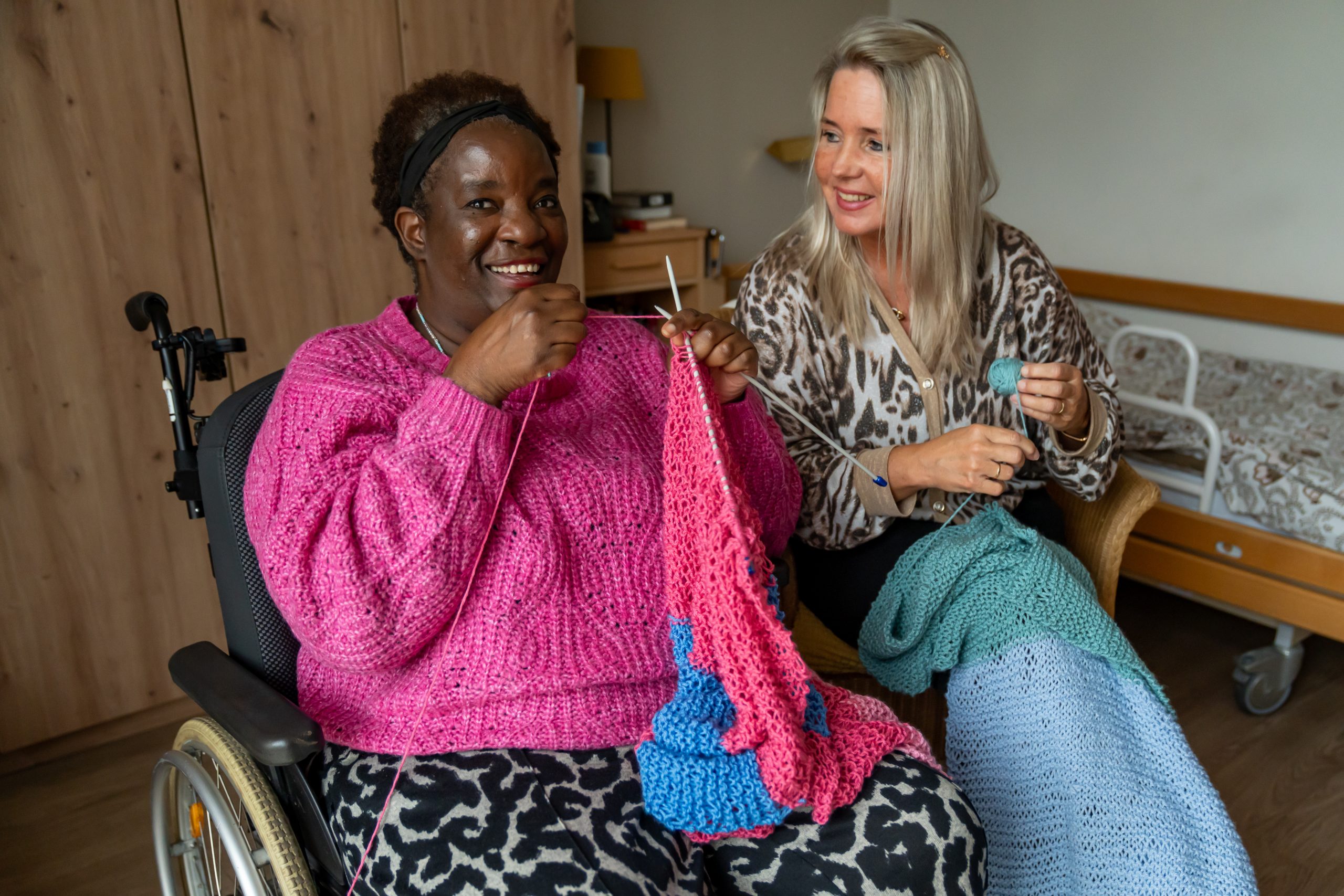 Twee vrouwen breien samen; een zit in een rolstoel, beiden lachen in een kamer.