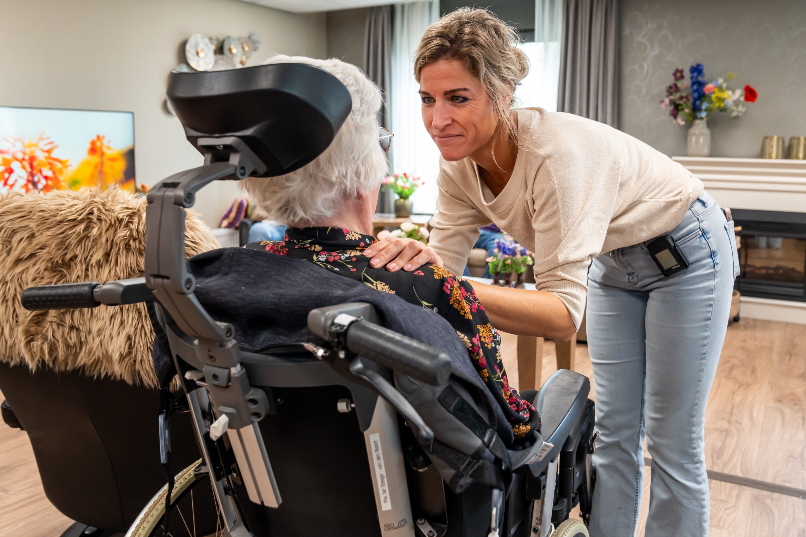 Verzorgende troost een oudere vrouw in een rolstoel in een huiskamer van een zorginstelling.
