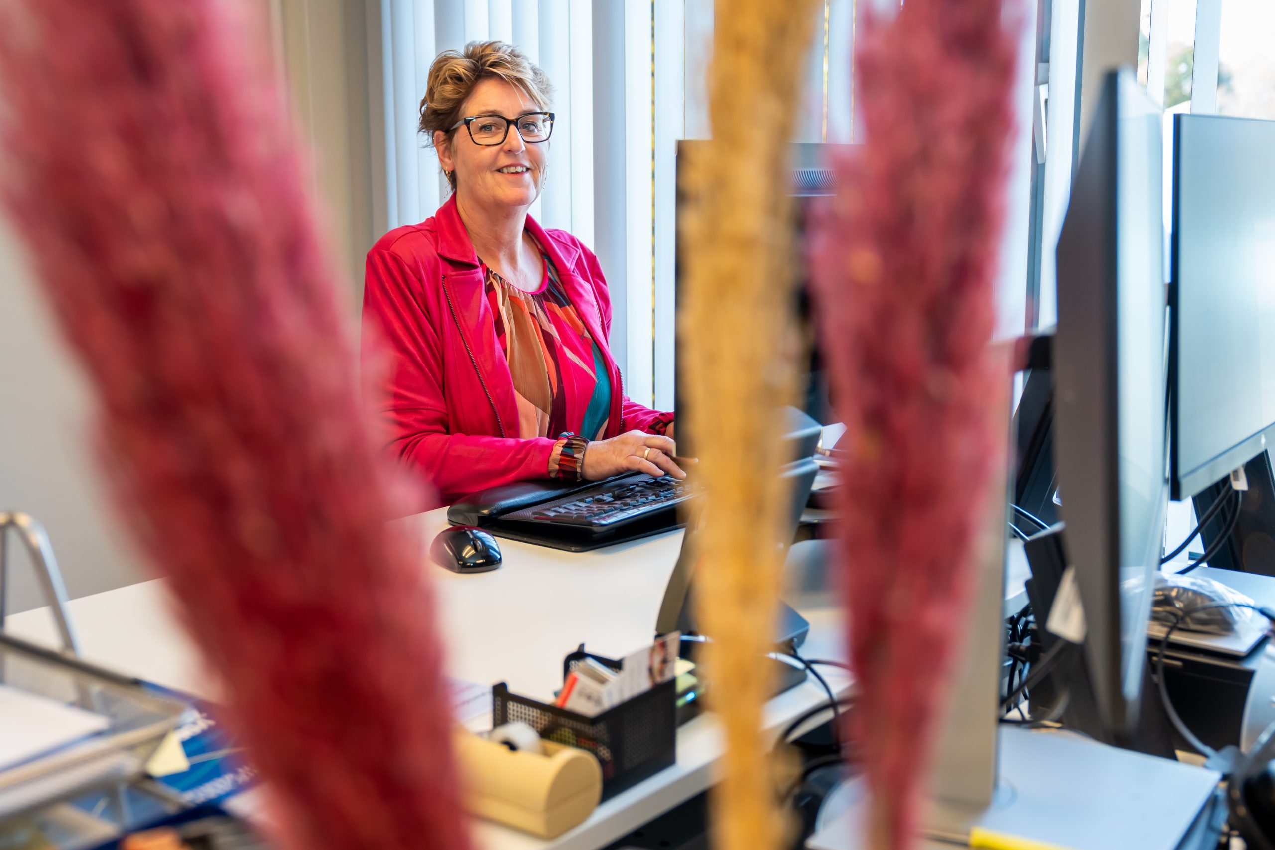 Vrouw met bril in roze jasje werkt glimlachend aan computer op kantoor, met onscherpe planten op de voorgrond.