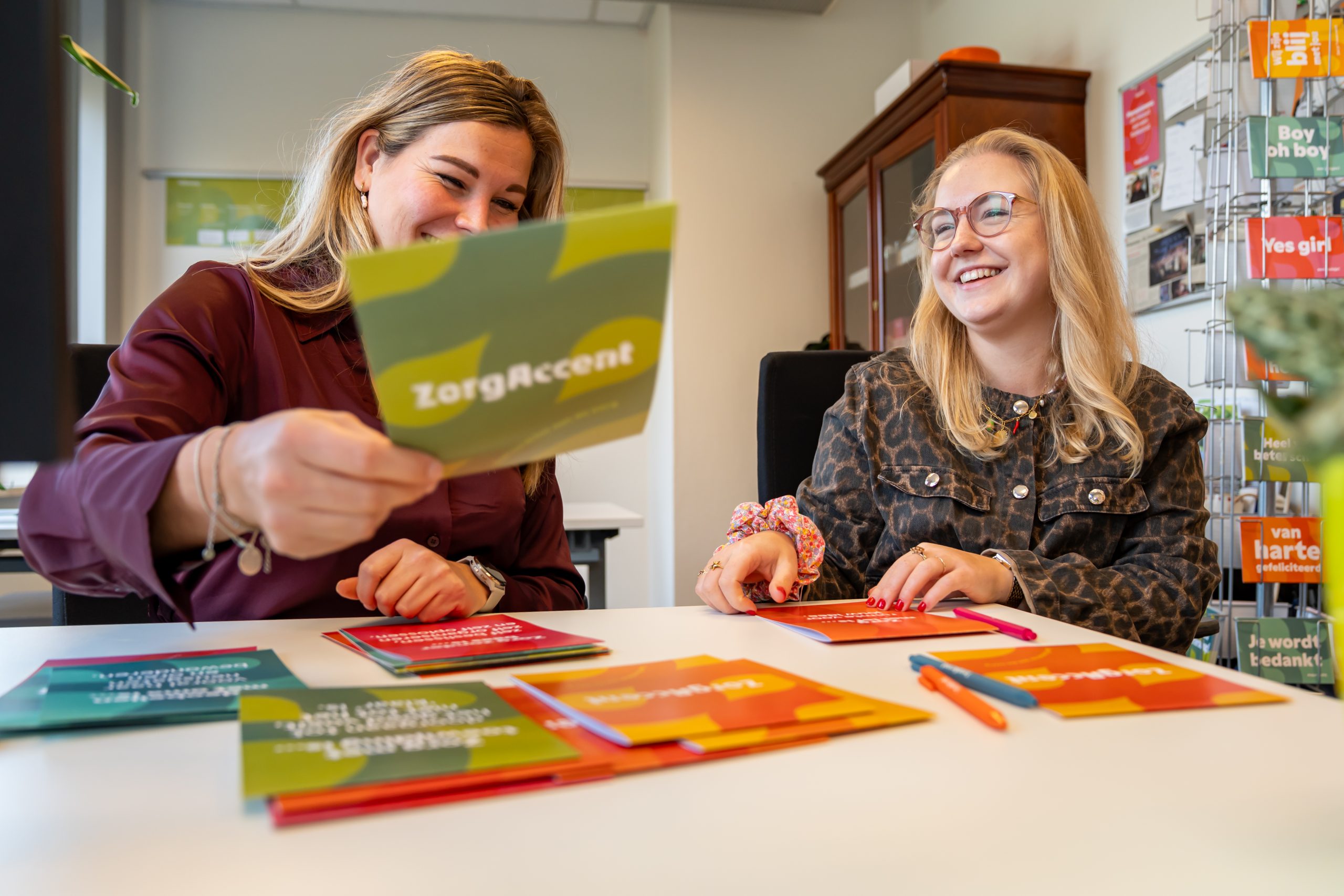 Twee collega's glimlachen terwijl ze kleurrijke ZorgAccent-kaarten sorteren aan een bureau.