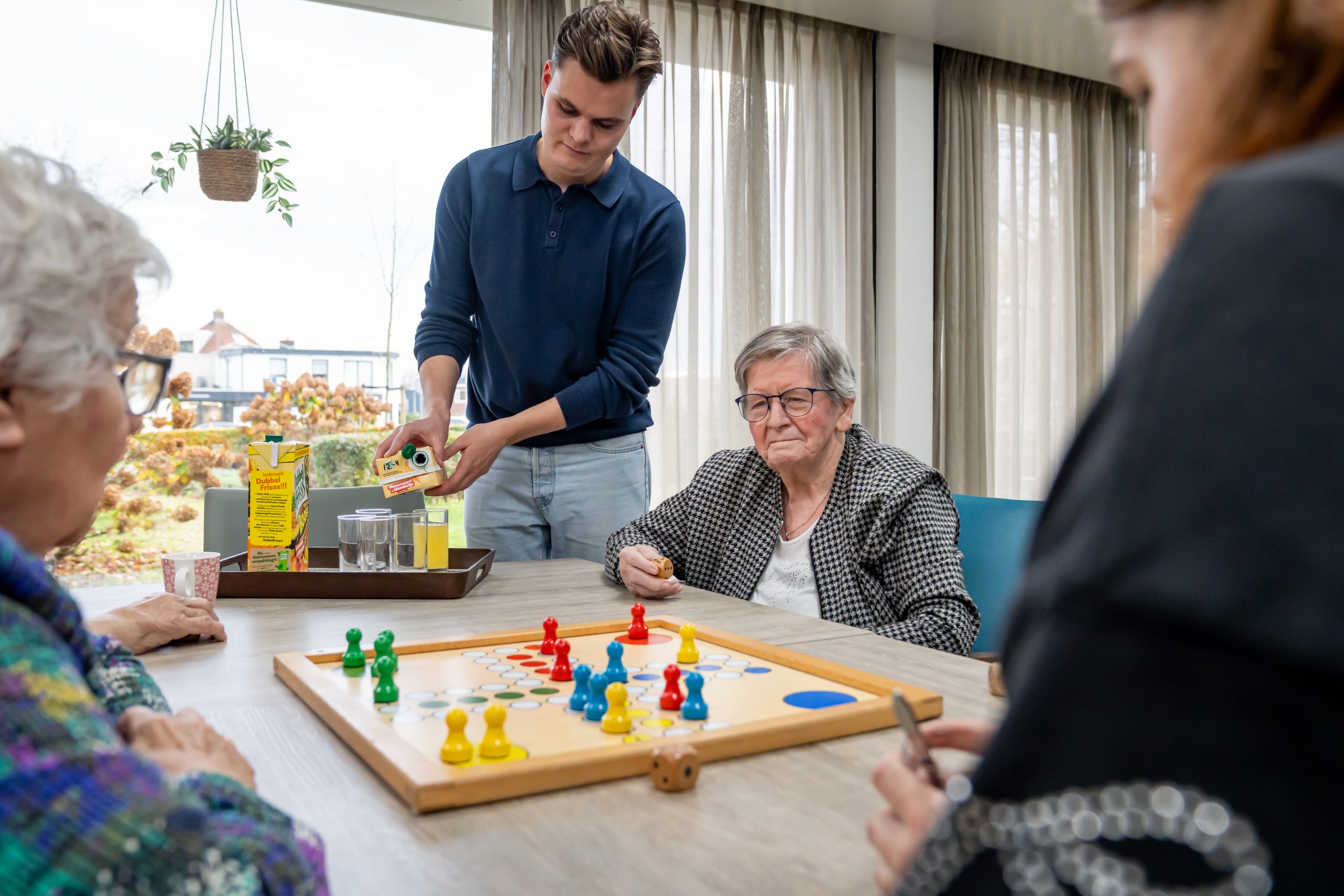 Jonge begeleider schenkt sap in tijdens een bordspel met ouderen aan tafel.
