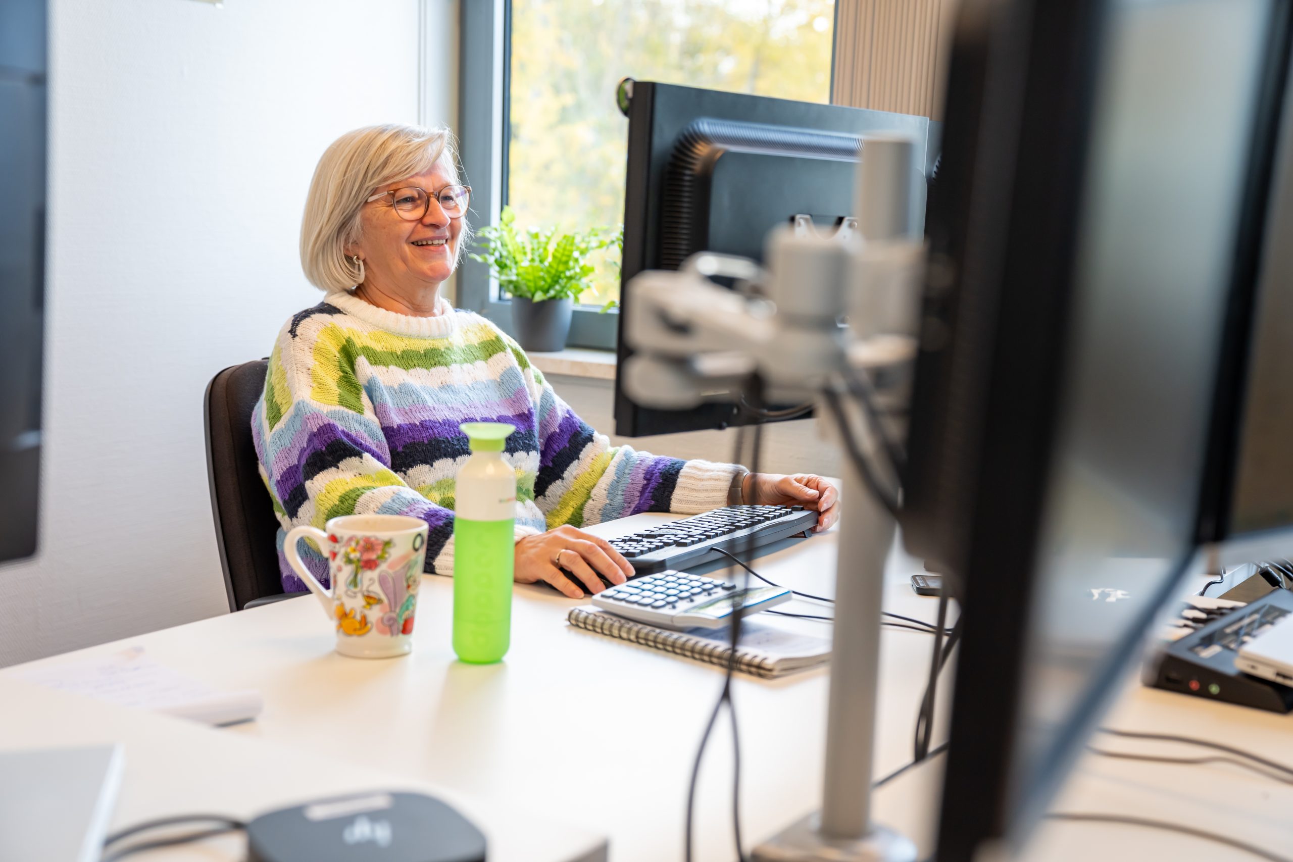 Glimlachende vrouw met bril werkt achter computer op kantoor met planten en mok.