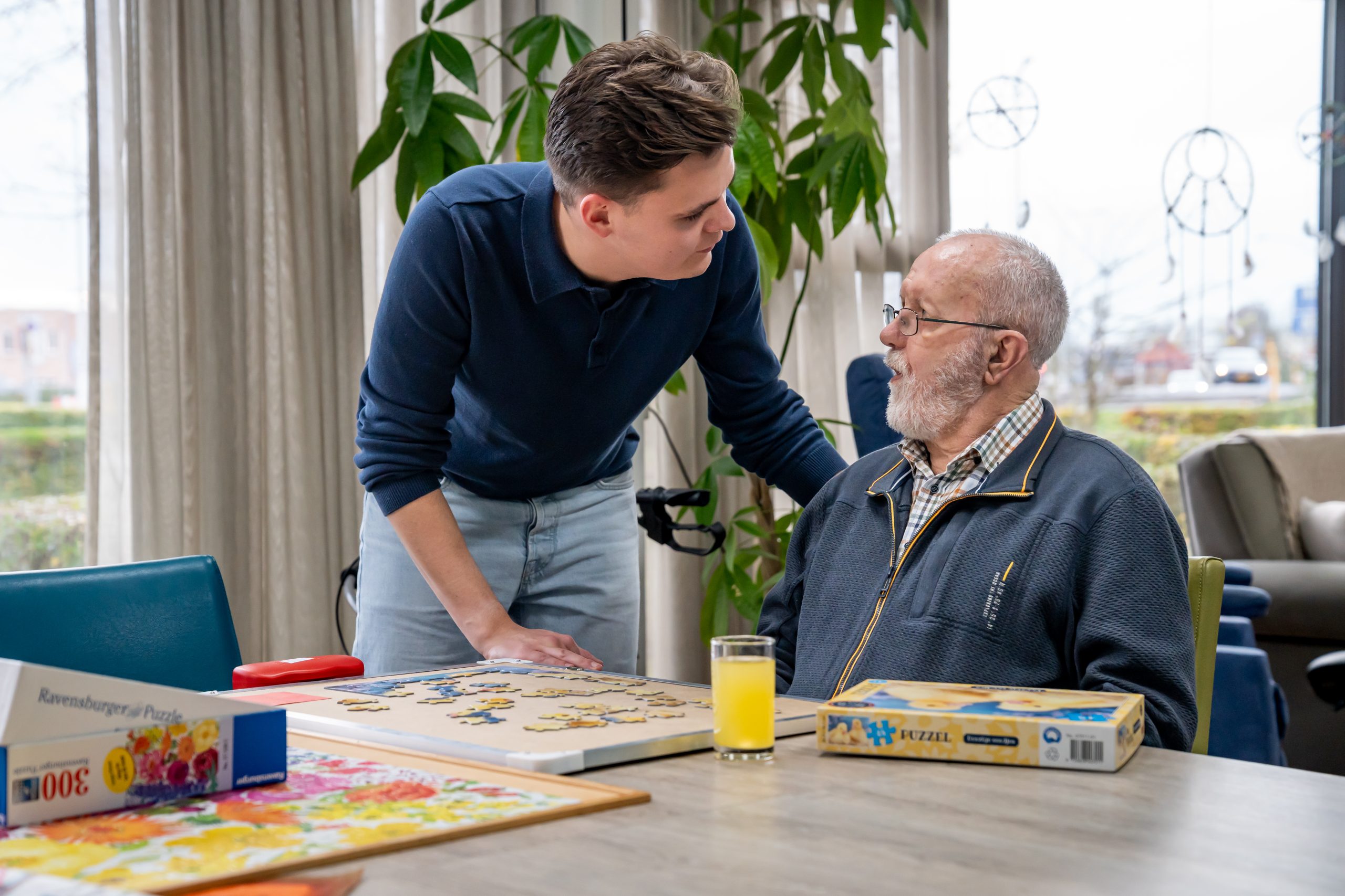 Jonge man helpt oudere man met een legpuzzel aan tafel in een lichte woonkamer.