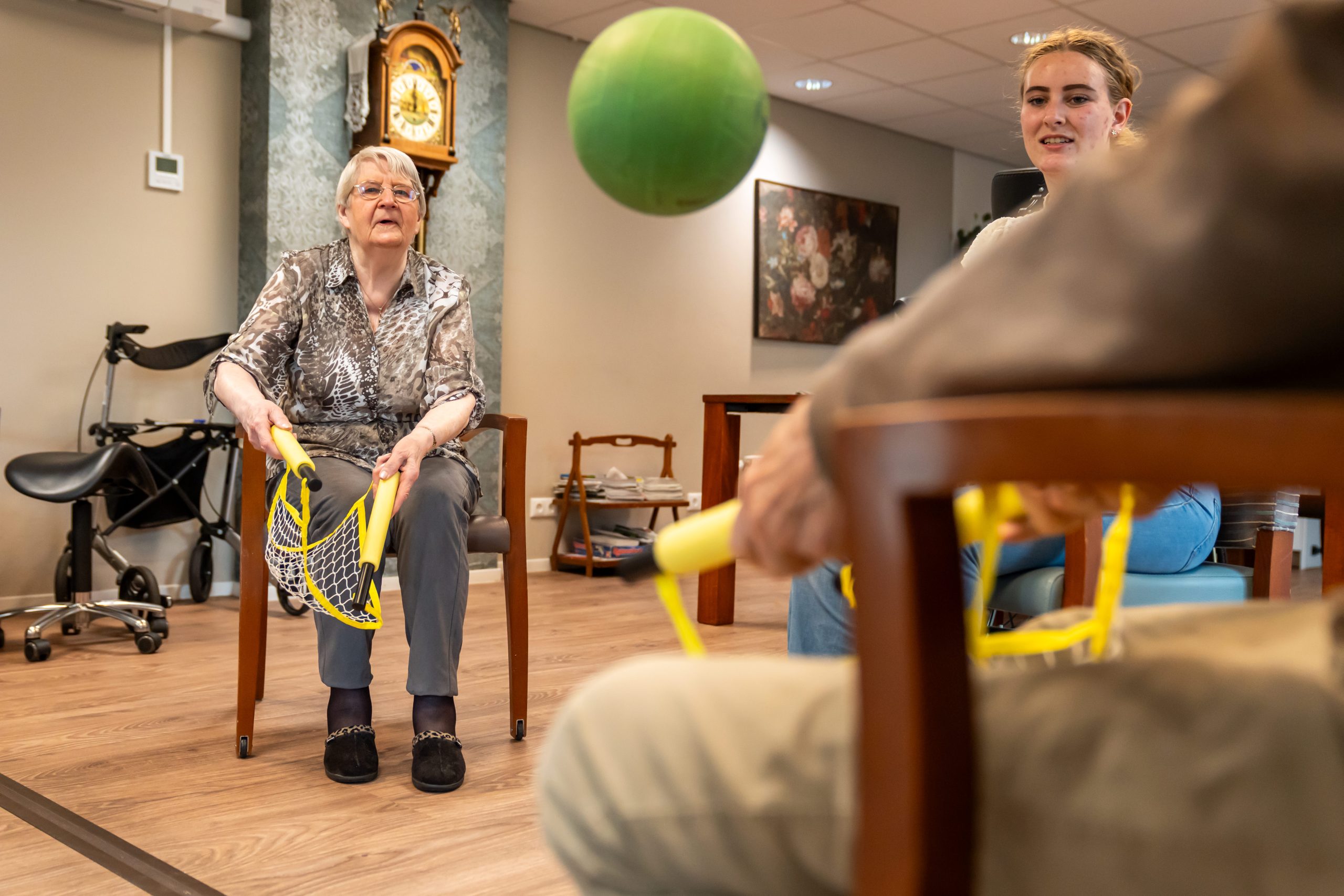 Ouderen spelen zittend een spel met vangnetten en een groene bal in een zorginstelling.