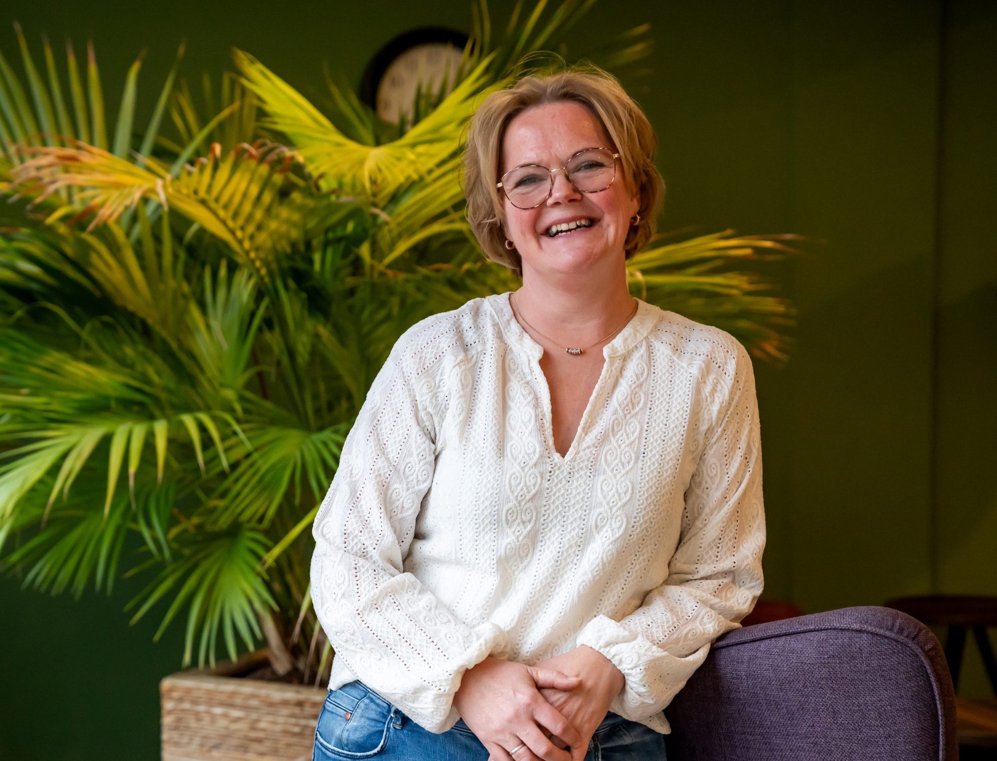 Vrolijke vrouw met bril in witte blouse, zittend voor groene kamerplant.