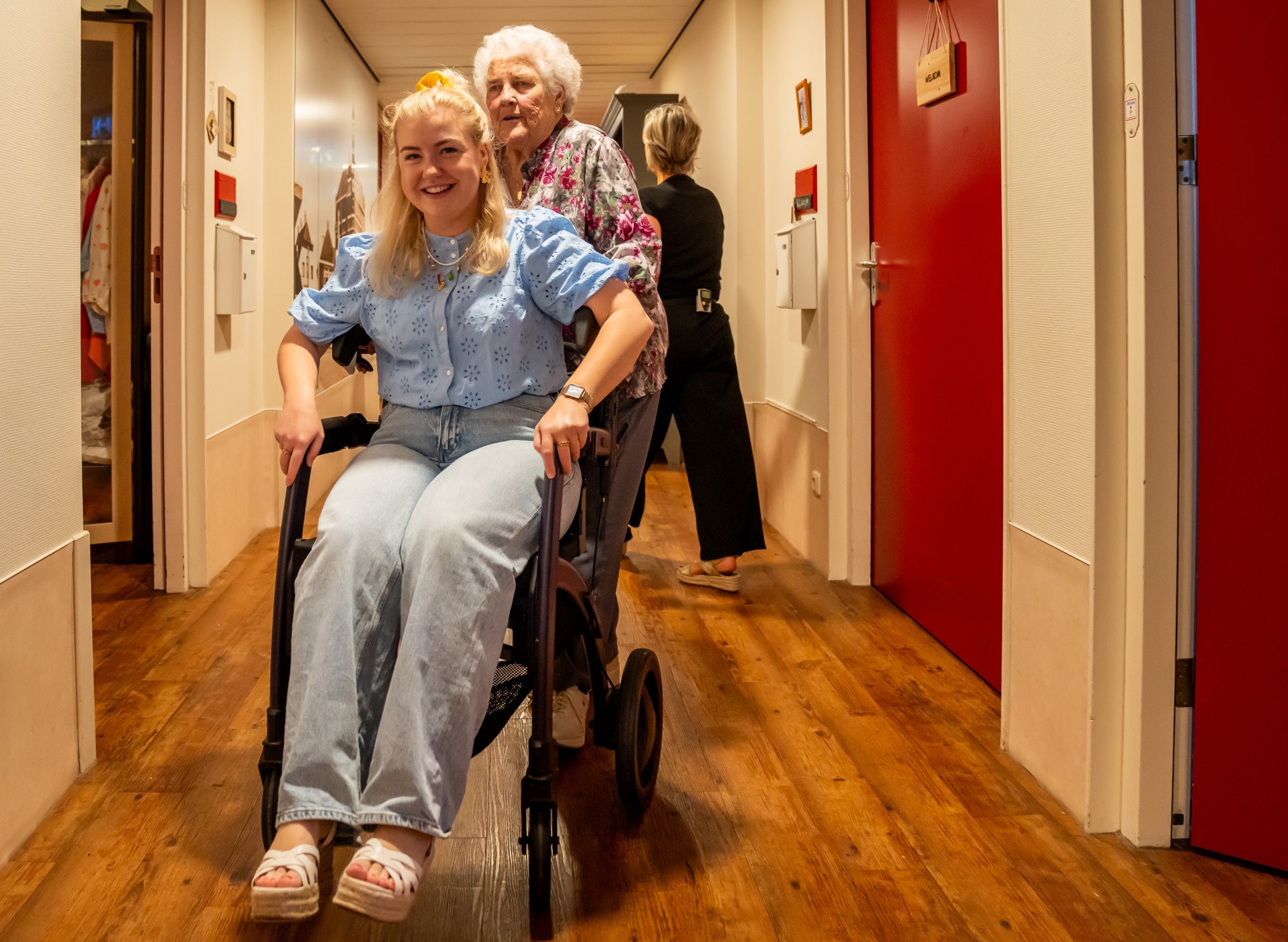 Jonge vrouw lacht in rolstoel terwijl oudere vrouw haar duwt in gang van zorginstelling.