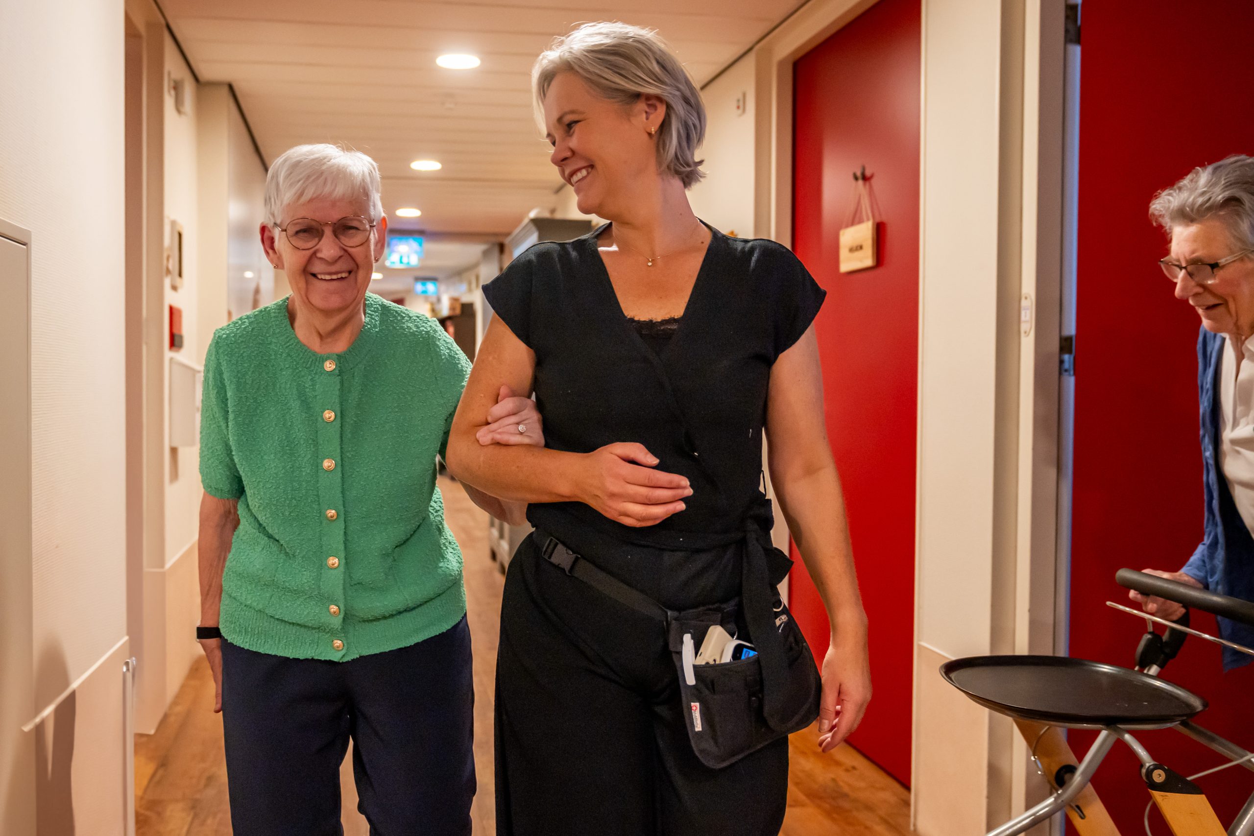 Zorgmedewerker loopt arm in arm met lachende oudere vrouw in een gang, met nog een mevrouw met rollator.