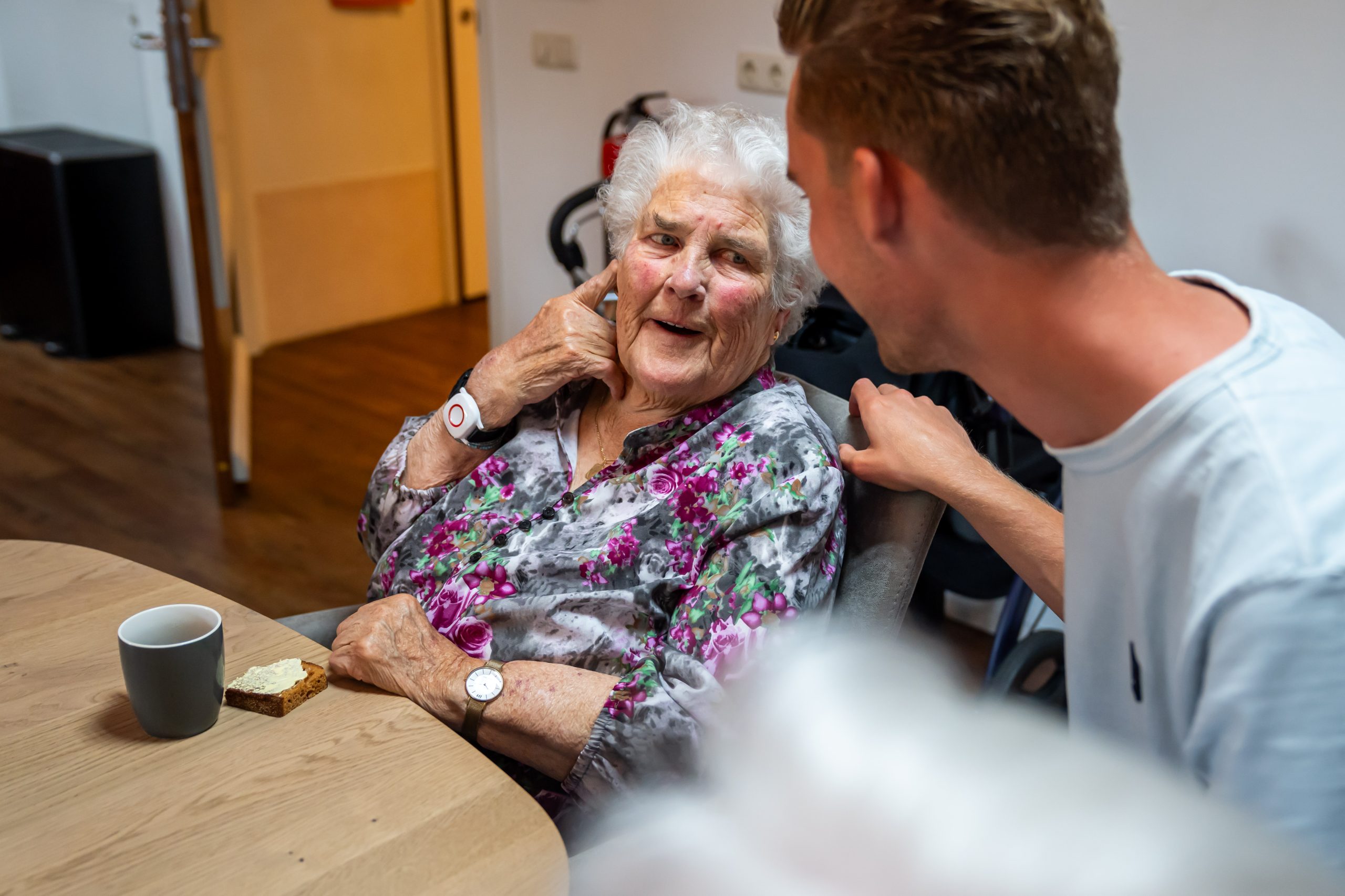 Oudere vrouw glimlacht en praat met jonge man aan tafel met koffie en brood.