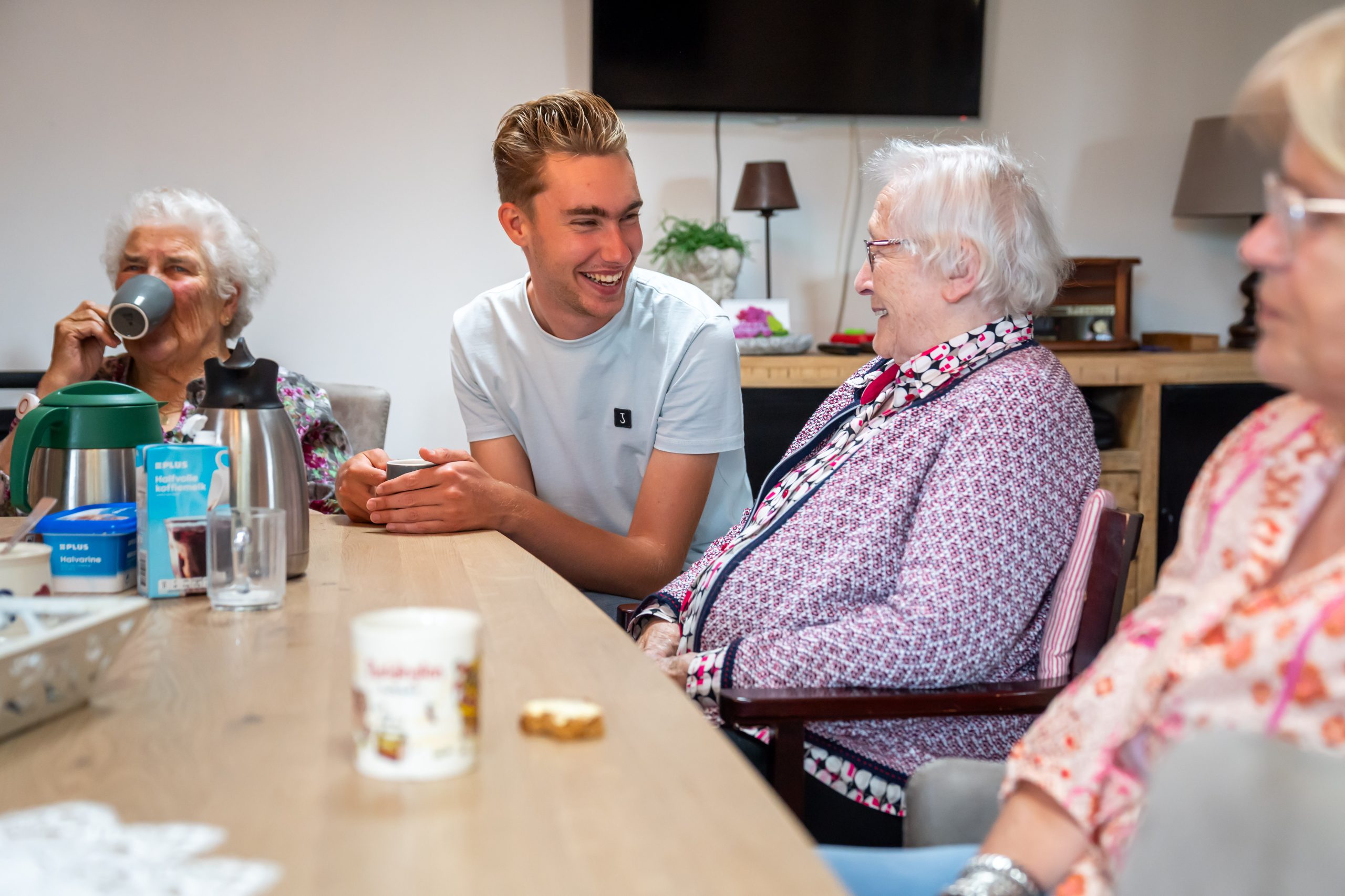Jonge man en ouderen lachen samen aan tafel met koffie en thee in een huiskamer.