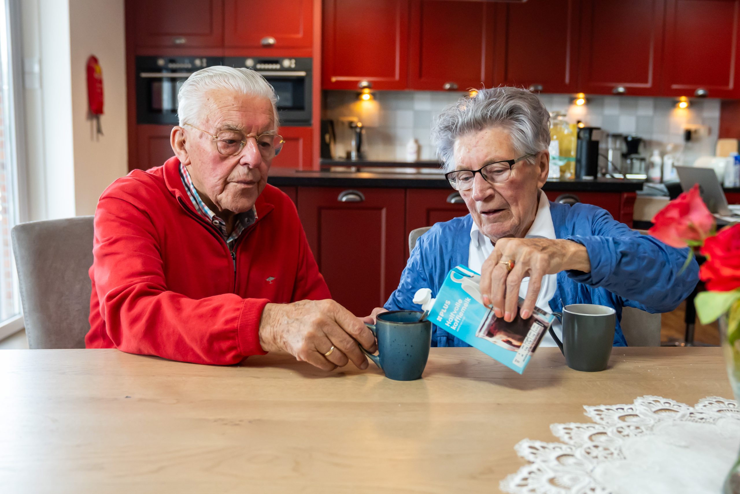 Twee ouderen aan tafel; vrouw giet melk in koffie terwijl man de mok vasthoudt, in keuken met rode kasten.