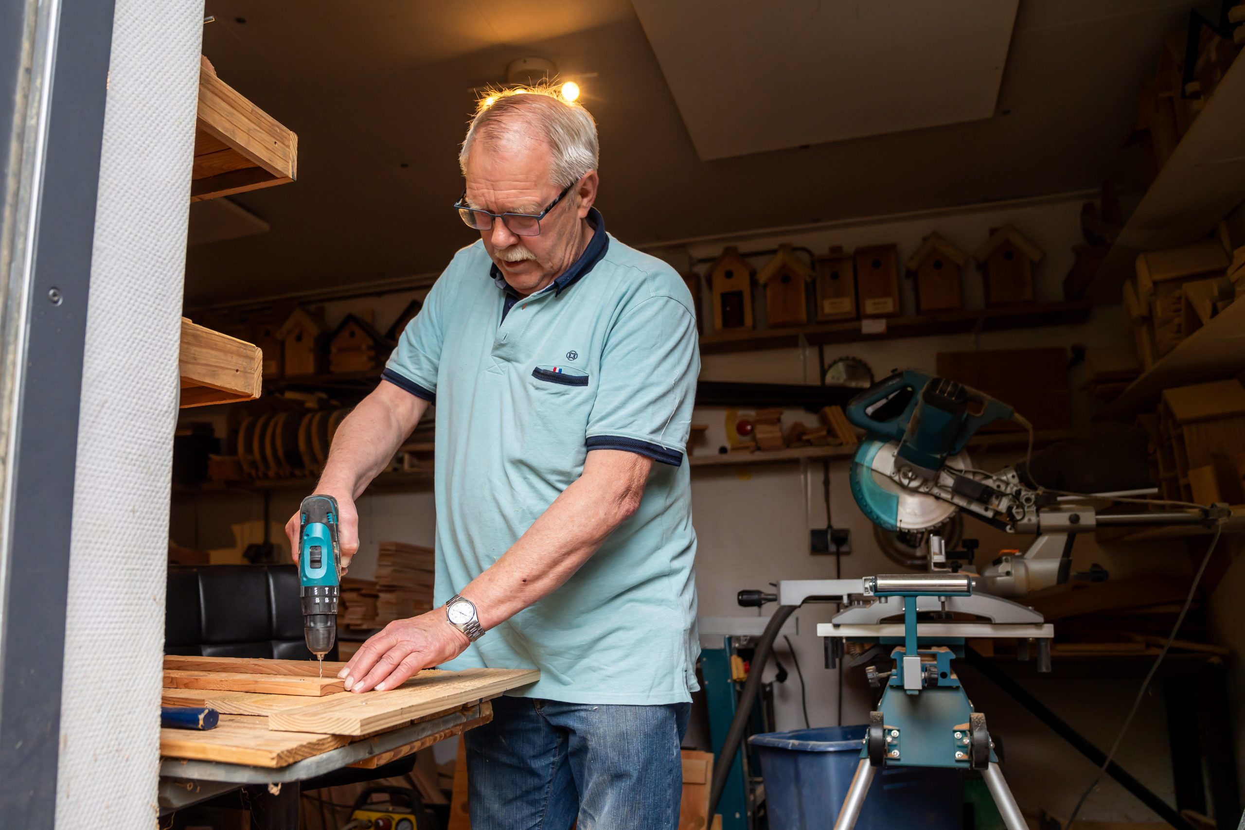 Man boort gaten in houten planken in een werkplaats met zaagmachine en vogelhuisjes op de achtergrond.
