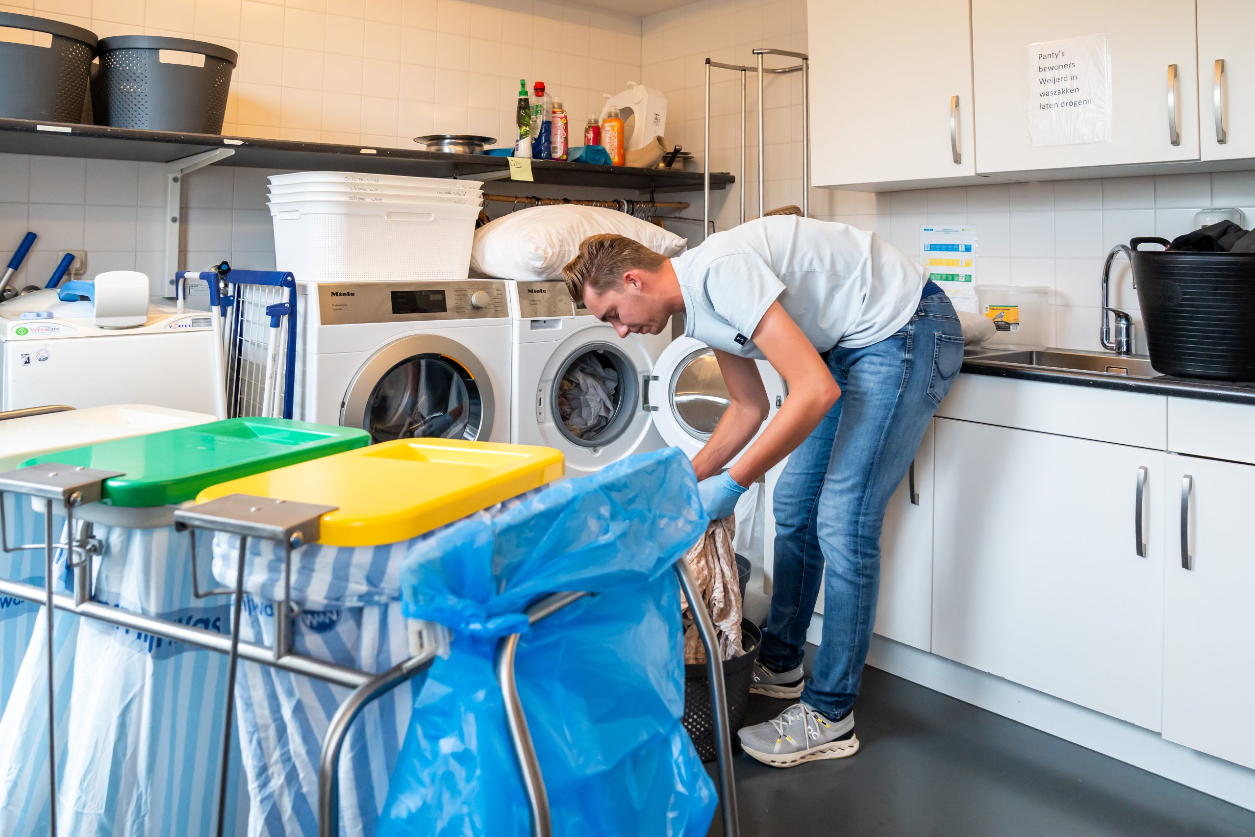 Medewerker laadt was in wasmachine in wasruimte met sorteertrolley en schoonmaakmiddelen.