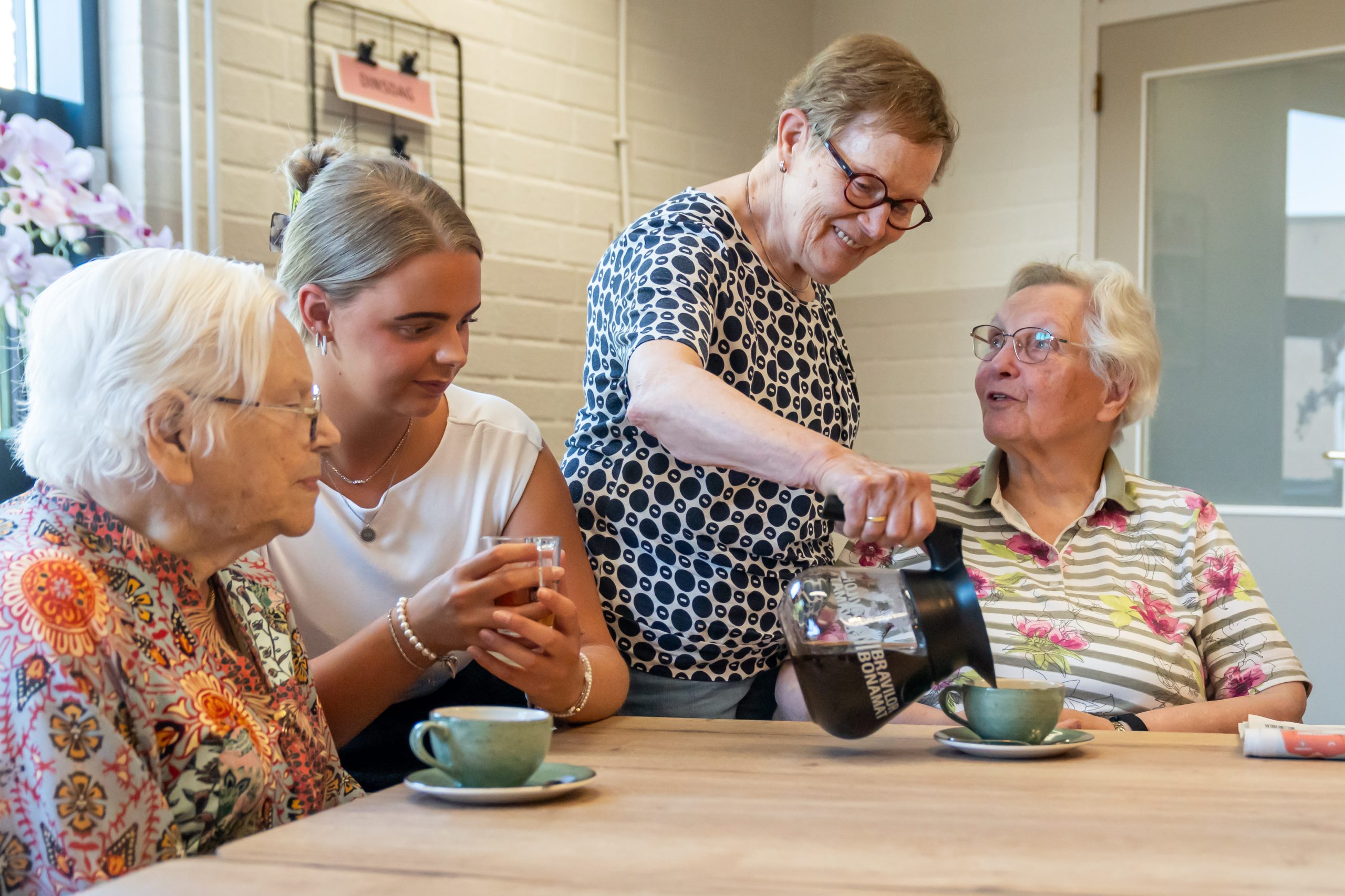 Vrouw schenkt koffie in voor een groep ouderen en een jonge vrouw aan tafel