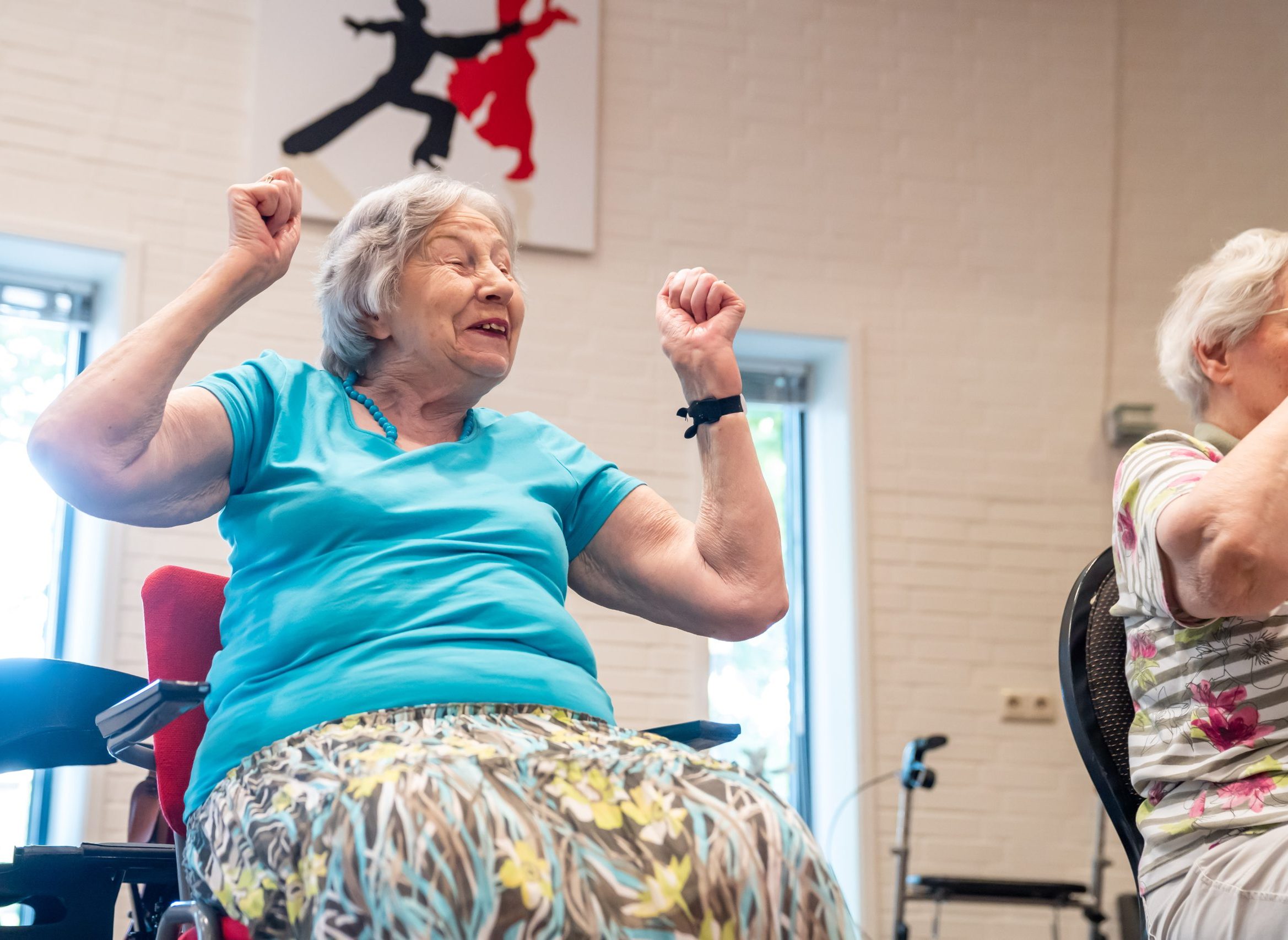 Oudere vrouw doet stoeloefeningen in een zaal, glimlachend met armen omhoog.