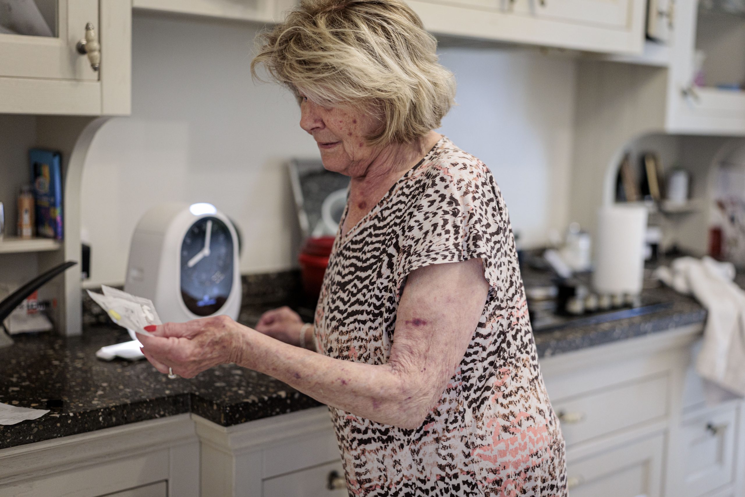 Oudere vrouw in keuken bekijkt een medicijnverpakking bij het aanrecht.