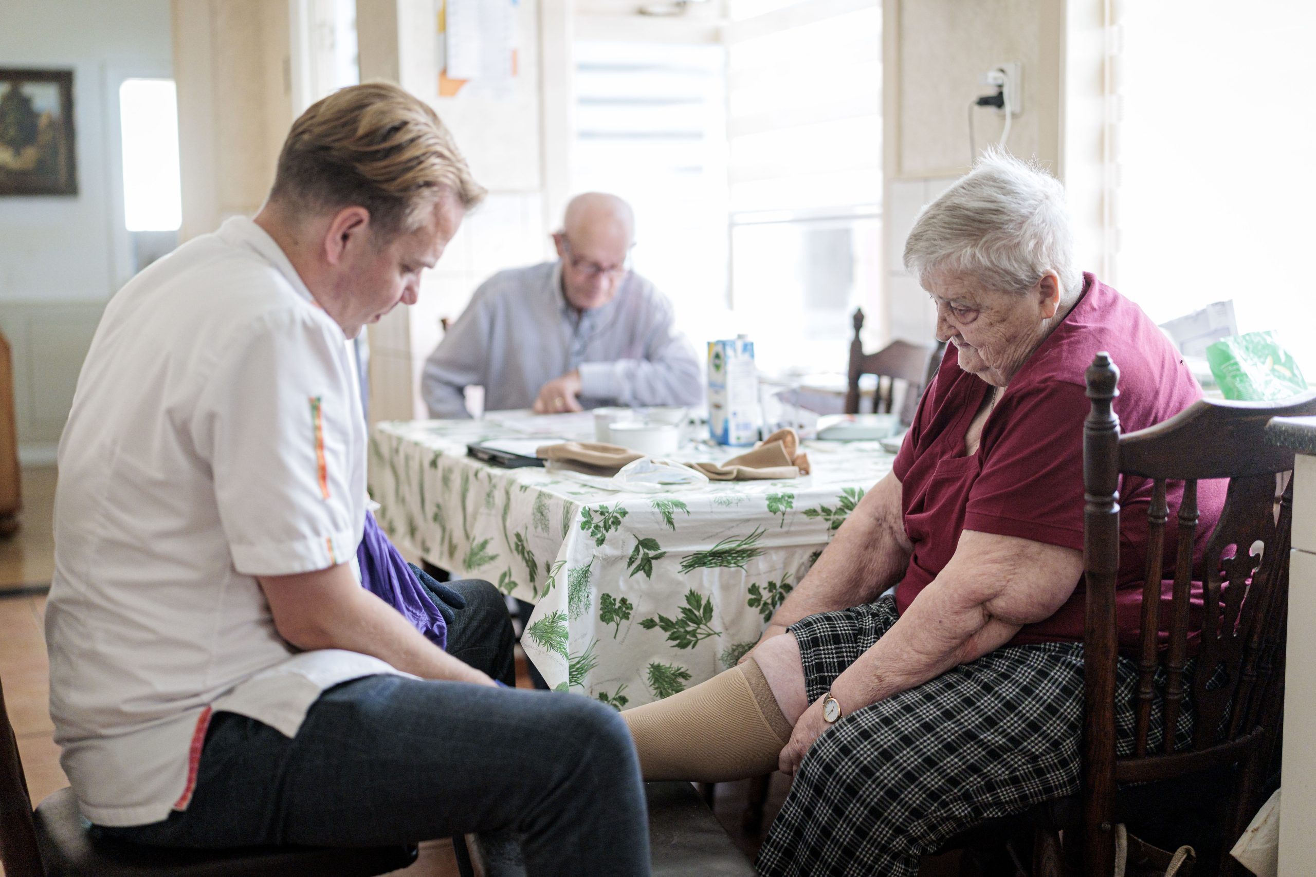 Zorgverlener helpt oudere vrouw met steunkous aan tafel, man leest op achtergrond.