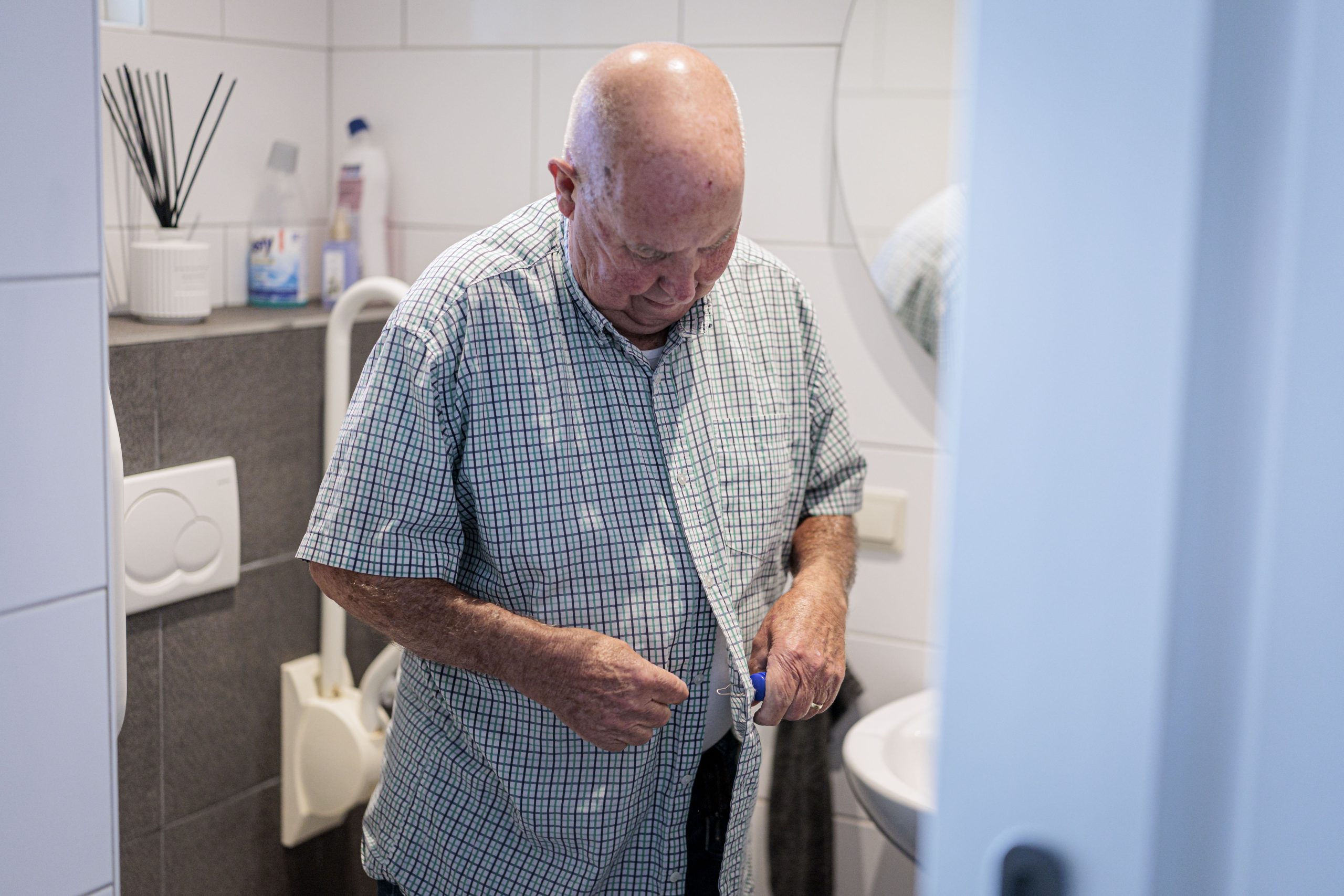 Oudere man knoopt zijn geruite overhemd dicht in een kleine betegelde badkamer.
