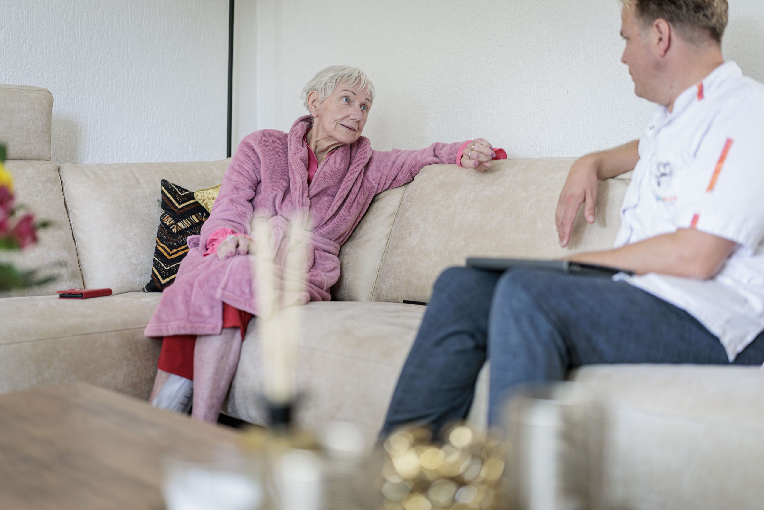 Oudere vrouw in roze badjas praat met zorgverlener op beige bank in woonkamer.