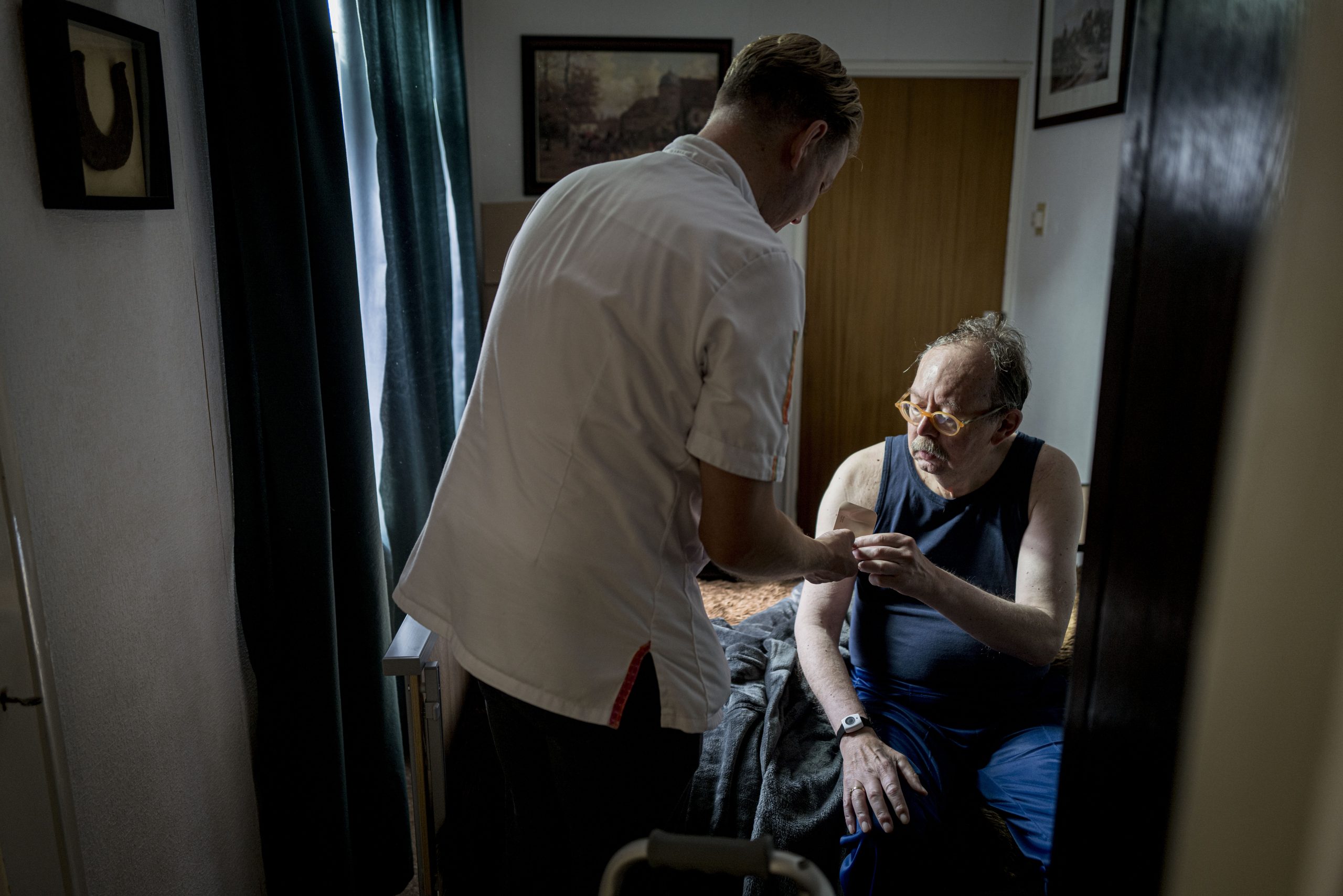Verzorger helpt zittende man in slaapkamer en reikt hem een voorwerp aan.