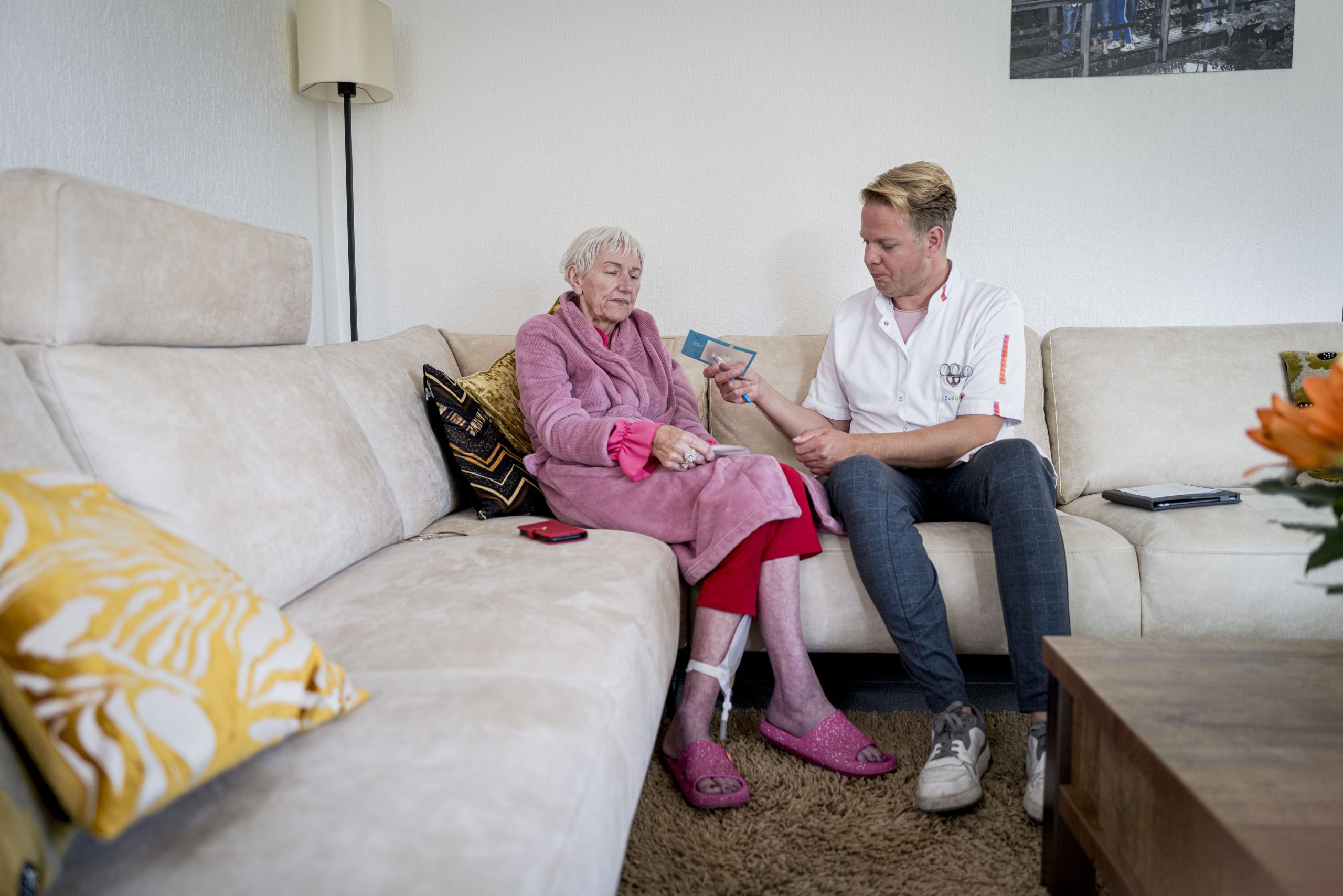 Thuiszorgverpleegkundige bespreekt medicatie met oudere vrouw in roze badjas op beige bank in woonkamer.