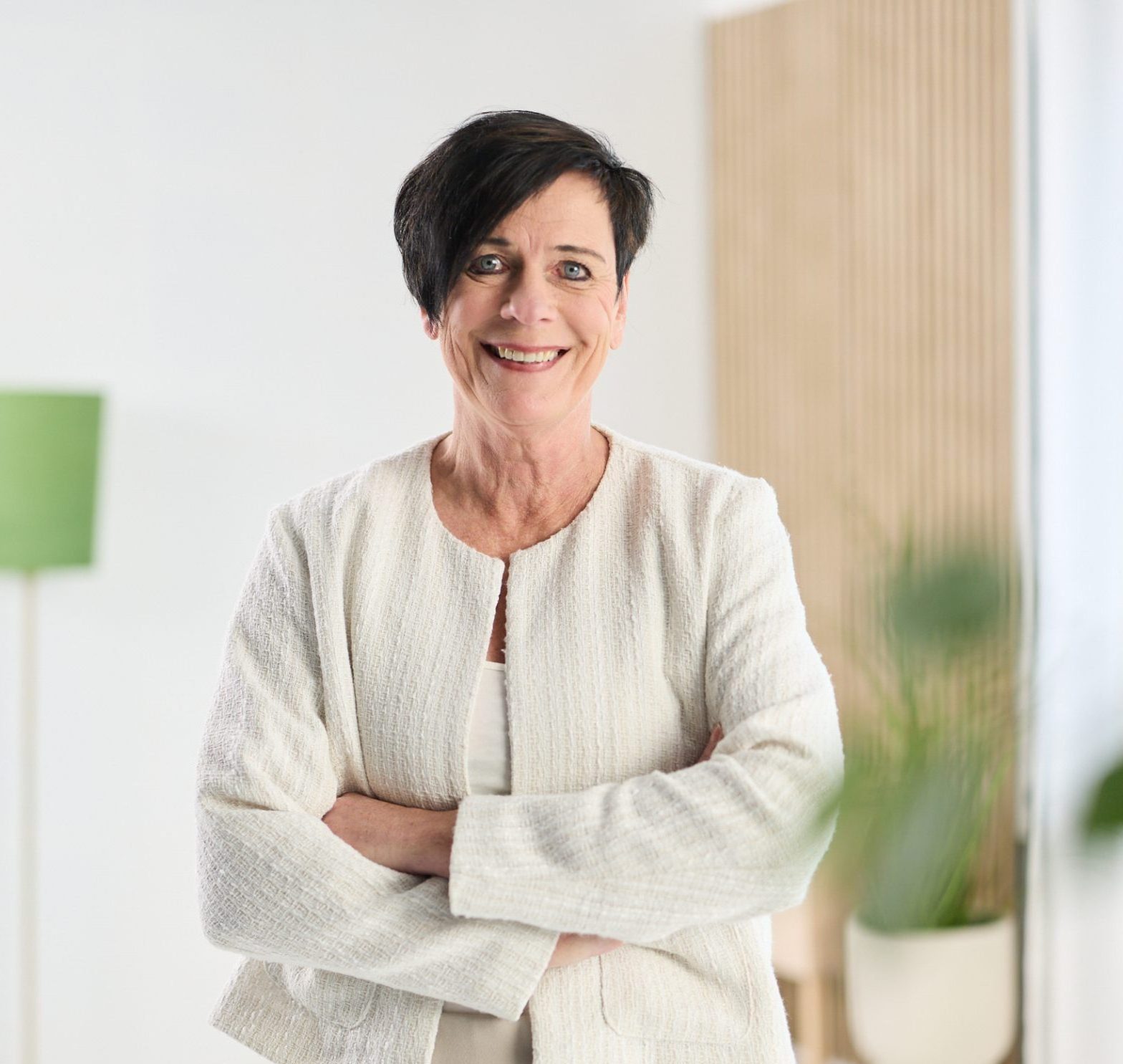 Lachende vrouw met kort donker haar in wit jasje, armen gekruist, in lichte kantoorruimte.