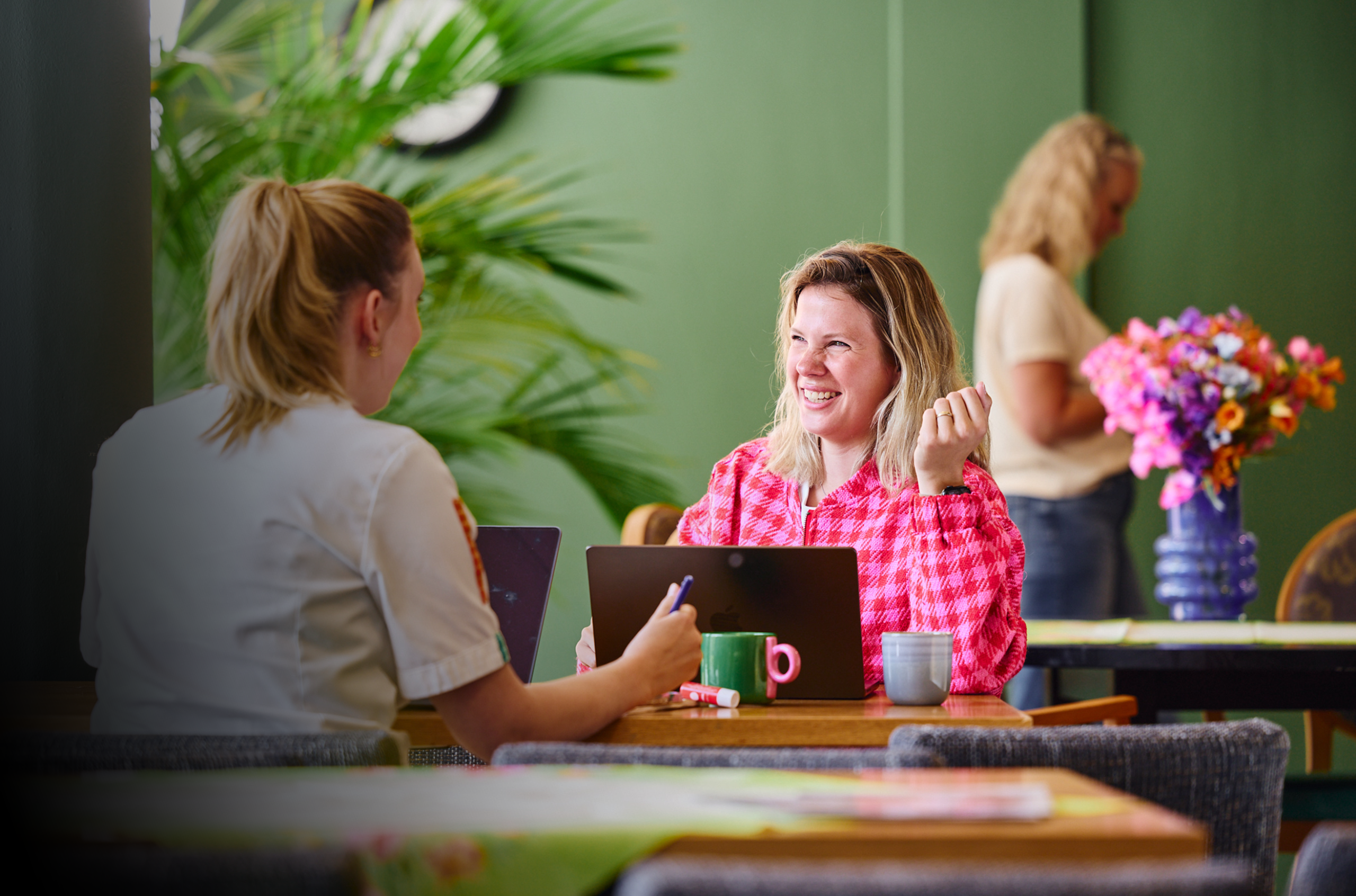 Twee vrouwen overleggen met laptops in een lichte, groene ruimte, met bloemenvaas en koffiemokken op tafel.