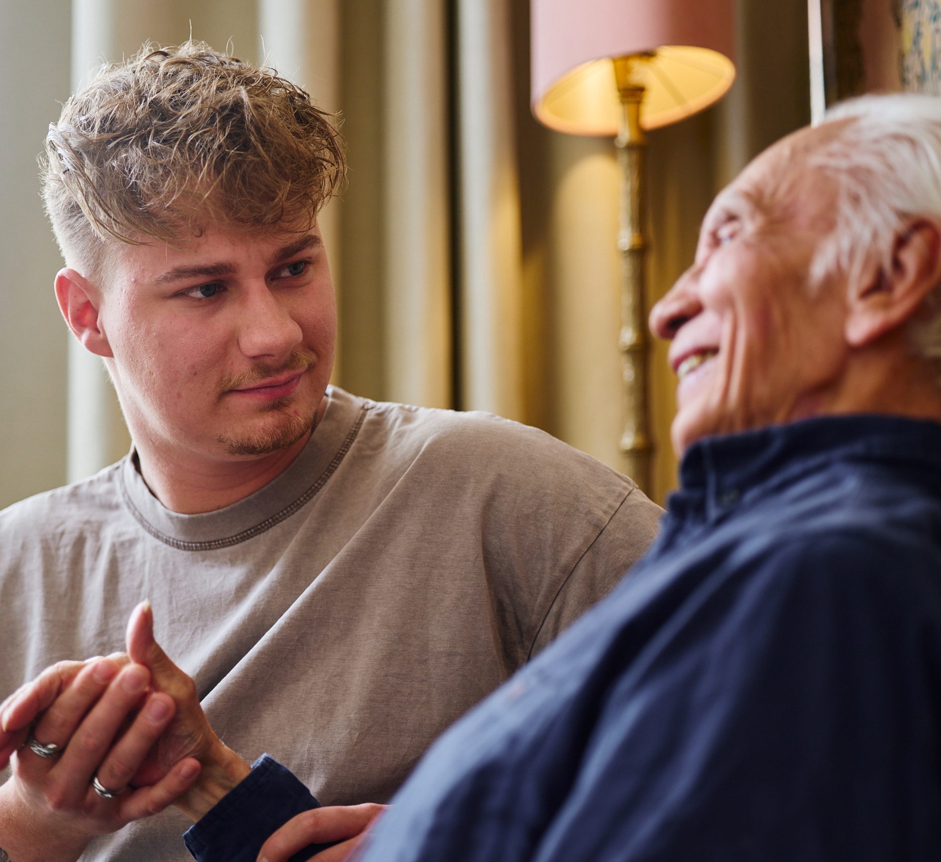 Jonge man houdt de hand van een oudere man vast en praat vriendelijk in een gezellige woonkamer.