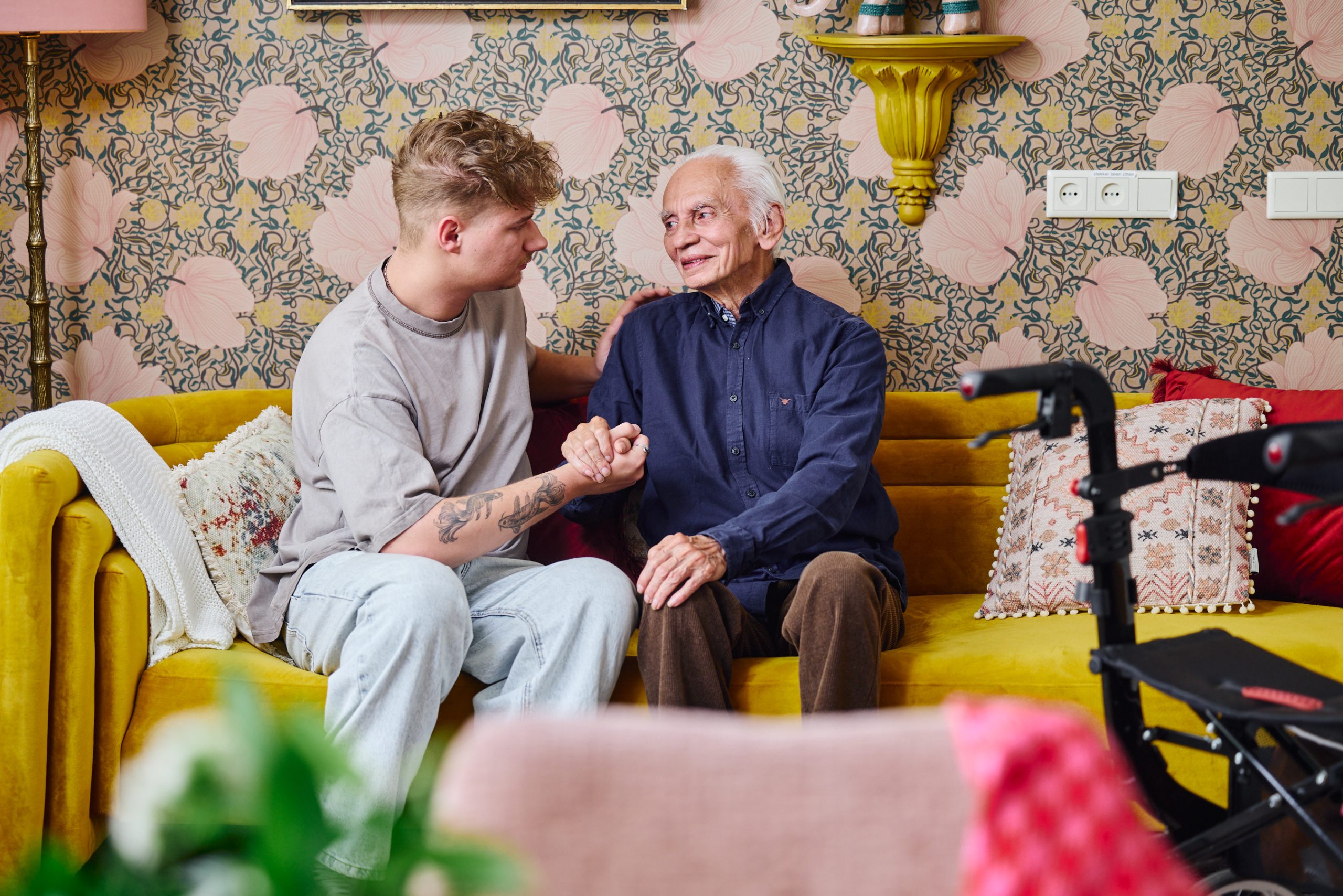 Jonge man troost oudere man op gele bank, hand in hand, rollator op de voorgrond.