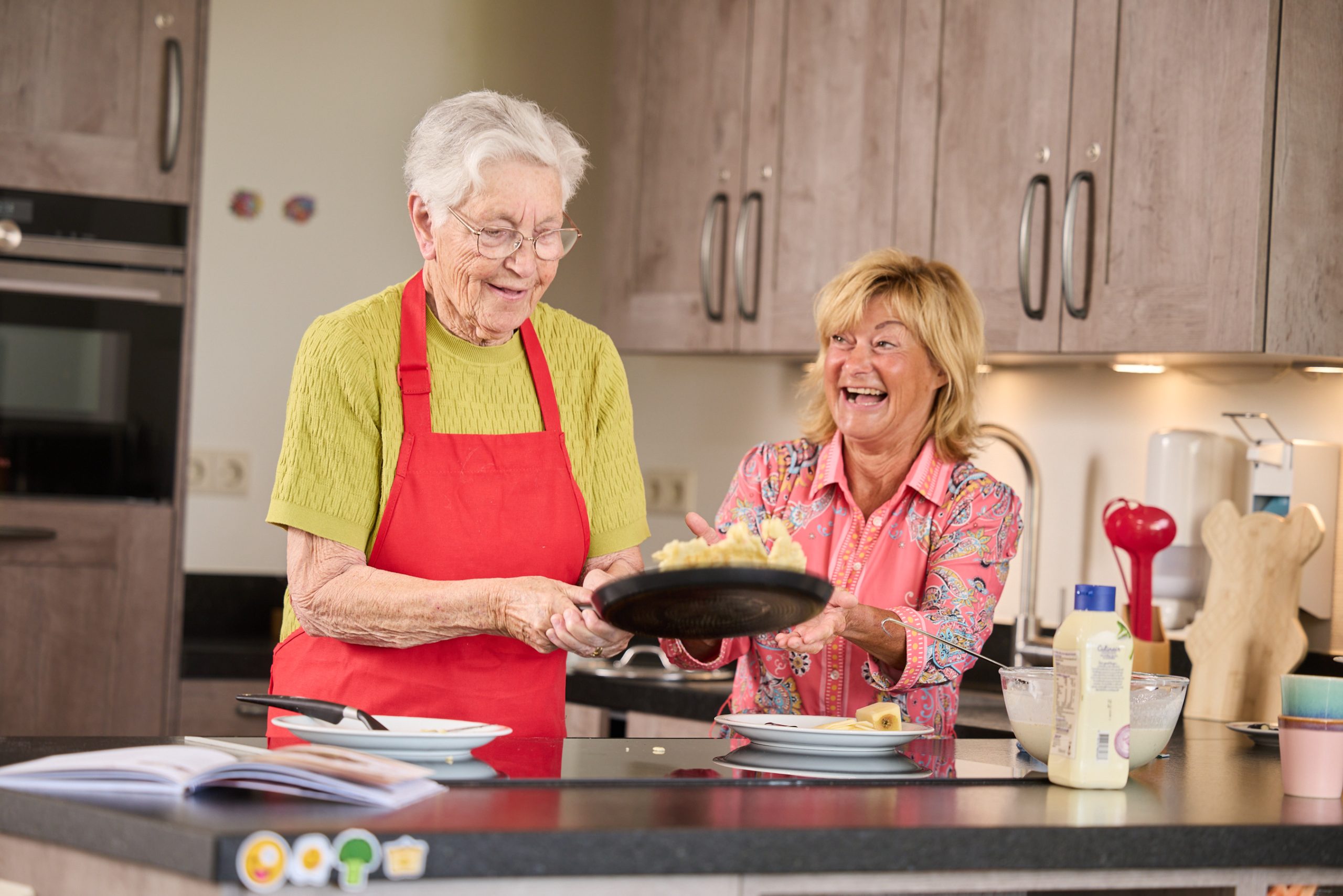 Twee vrouwen bakken en serveren pannenkoeken in een gezellige keuken, lachend samen.