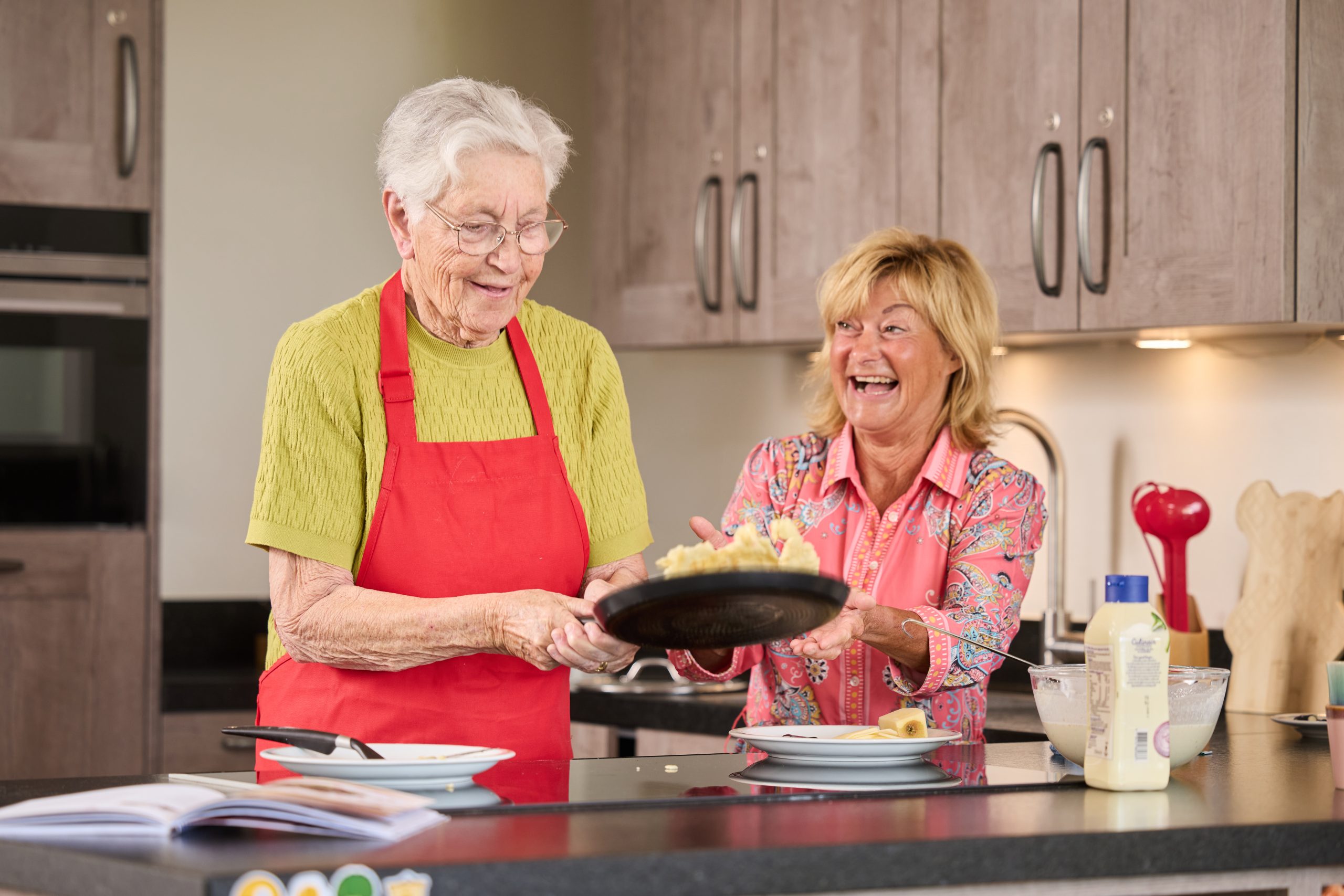 Oudere vrouw in rode schort bakt pannenkoeken met lachende vriendin in keuken.
