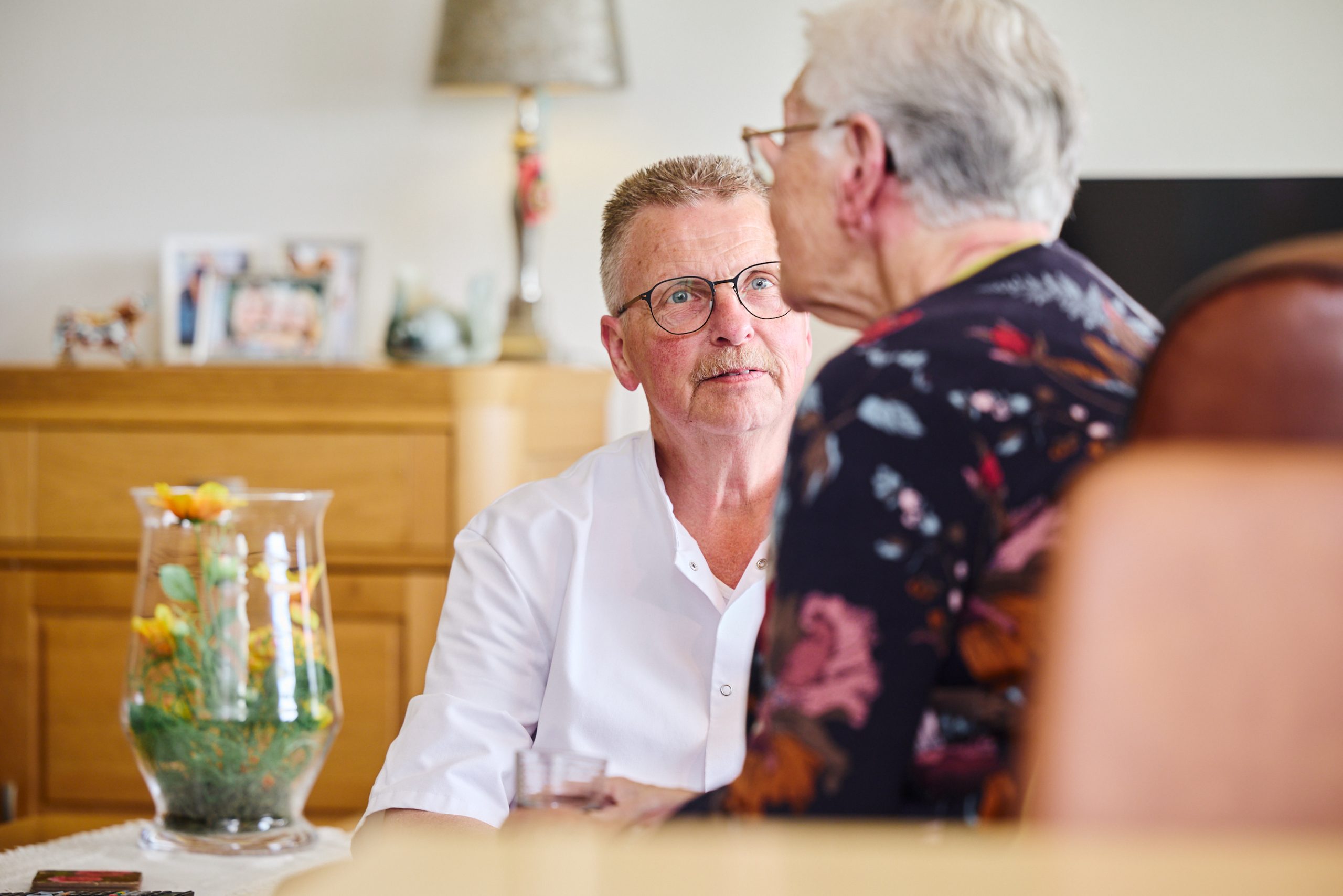 Zorgverlener praat met oudere vrouw in een warme woonkamer, naast een vaas met bloemen.