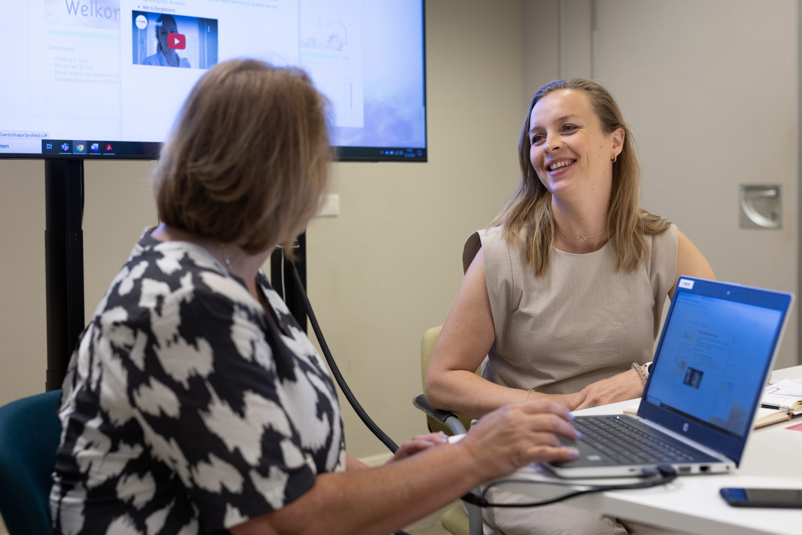 Twee collega’s praten lachend tijdens overleg met laptop en presentatiescherm op kantoor.